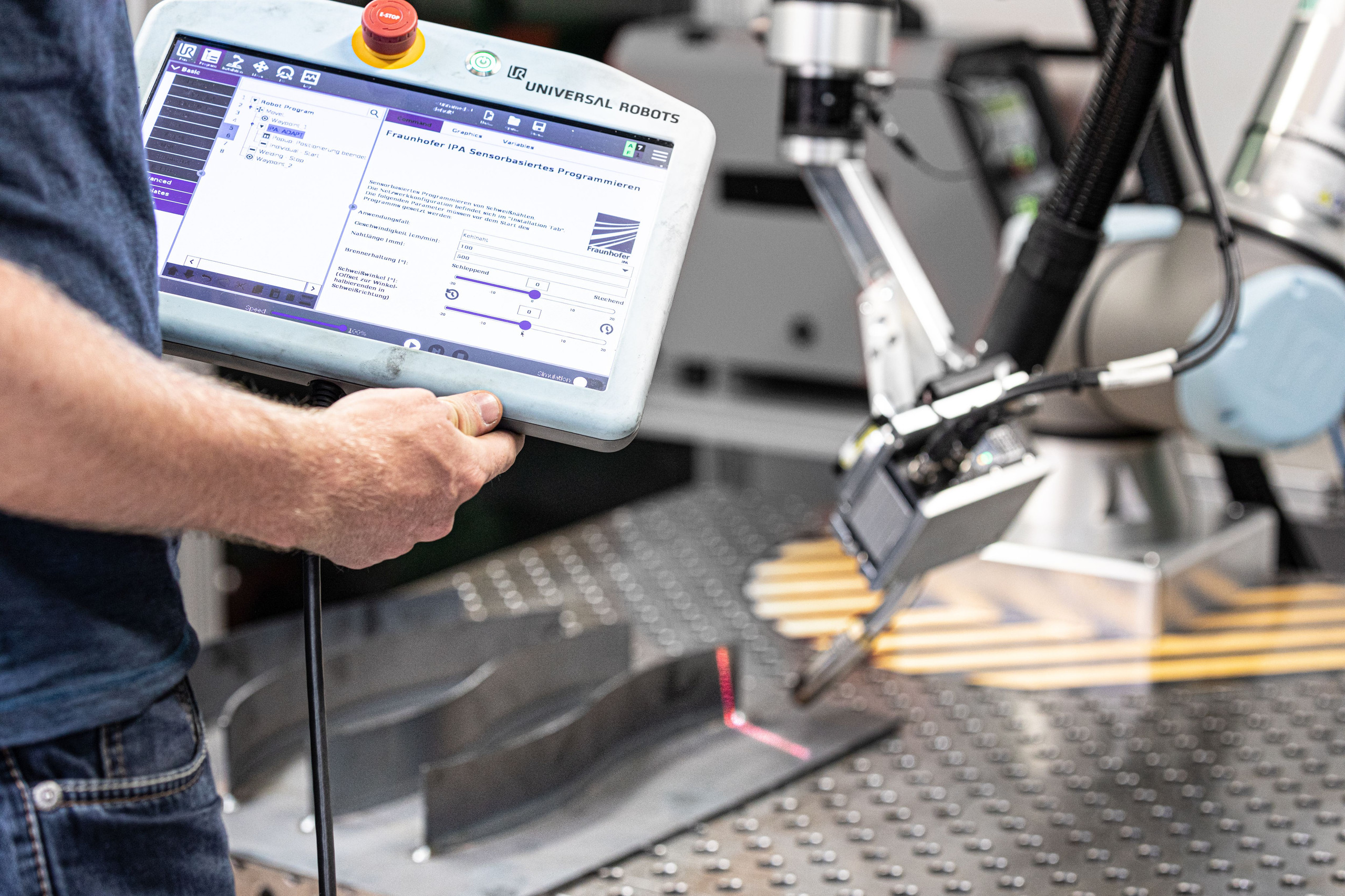 Schweißen Teilansicht eines Mannes von hinten. Er trägt ein Tablet. Vor ihm ein Werkstück aus Metall, auf dem eine Linie für die Schweißnaht zu sehen ist. Darüber ein automatisiertes Schweißgerät.