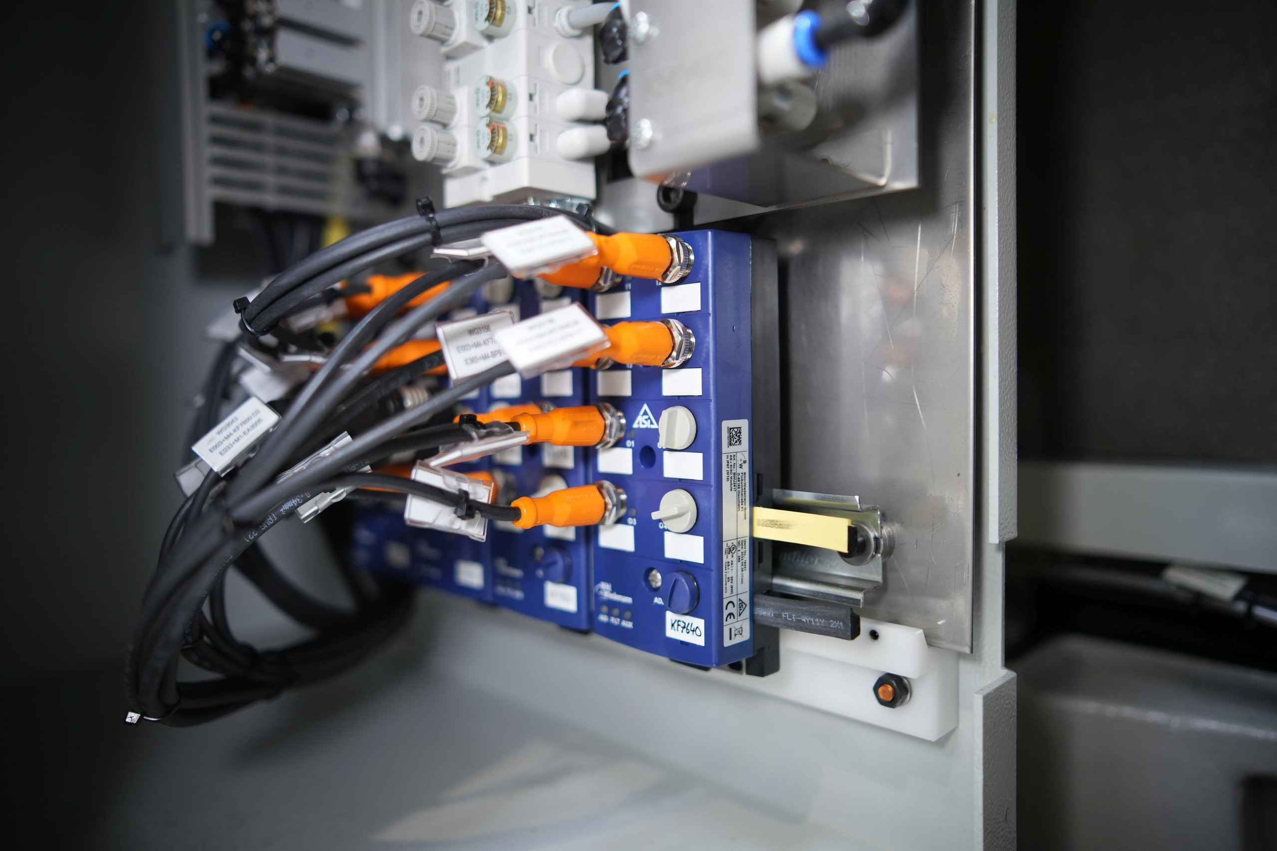 <p>Das Bild zeigt einen Ausschnitt aus dem Inneren eines Geräts oder einer Maschine, in dem sich mehrere elektronische Komponenten befinden. Im Mittelpunkt steht eine blaue Platine mit orangefarbenen Steckverbindungen. Um die Platine herum verlaufen Kabelstränge, die mit verschiedenen Anschlüssen verbunden sind. Es gibt auch Beschriftungen und möglicherweise Schalter oder Drehregler auf der Platine. Im Hintergrund sind weitere elektronische Elemente sichtbar, die Teil des Steuerungssystems sein könnten.<p>