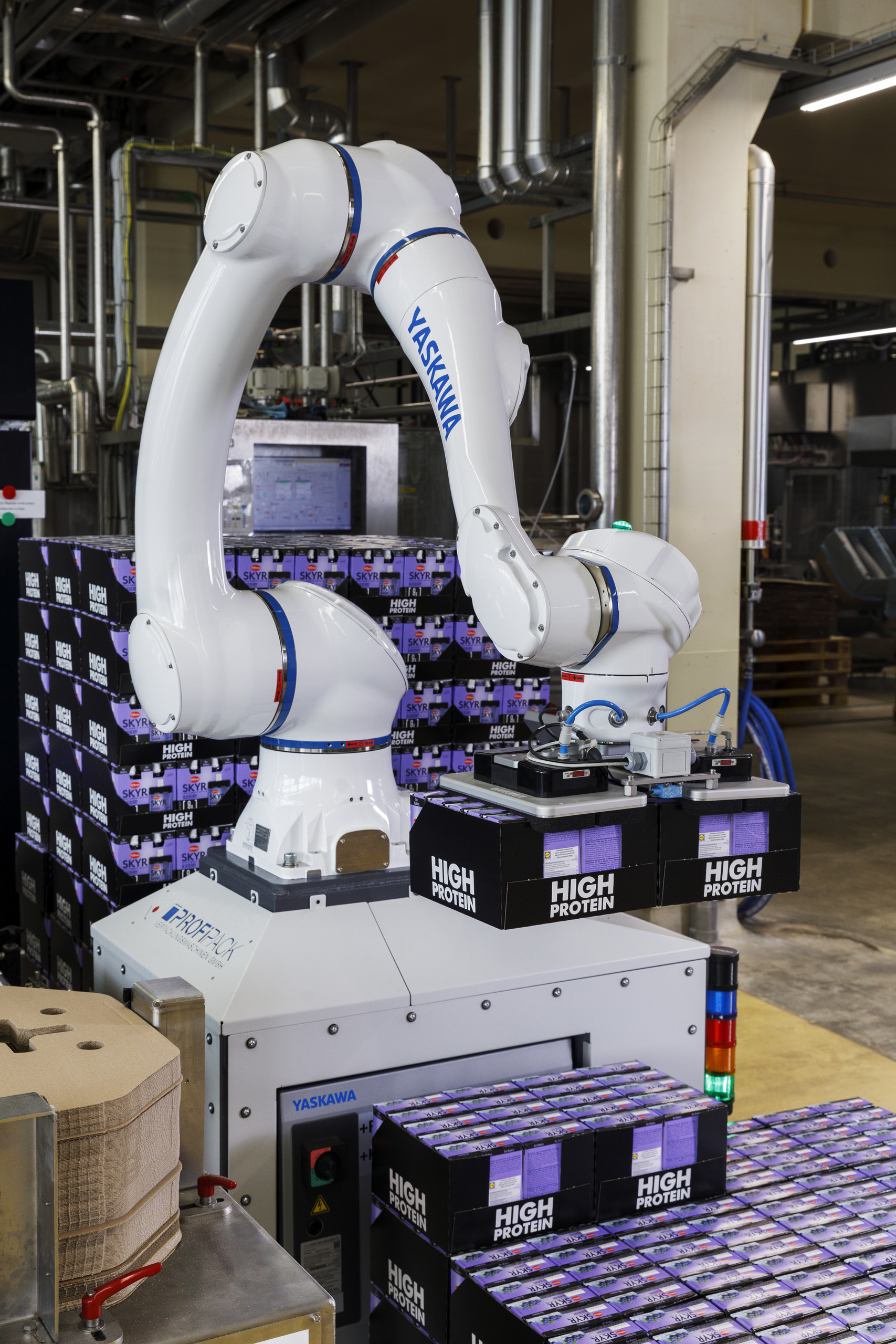 <p>Man sieht: Ein weißer Yaskawa-Cobot (Modell HC20DTP) in der Produktionshalle der Privatmolkerei Bauer. Der Roboterarm hebt mit einem Vakuumgreifer zwei schwarze Trays mit „High Protein“-Produkten an, um sie auf einer Palette zu stapeln. Im Vordergrund sind Karton-Zwischenlagen und bereits fertig gestapelte Joghurt-Gebinde auf der Anlage von Profipack zu sehen.<p>
