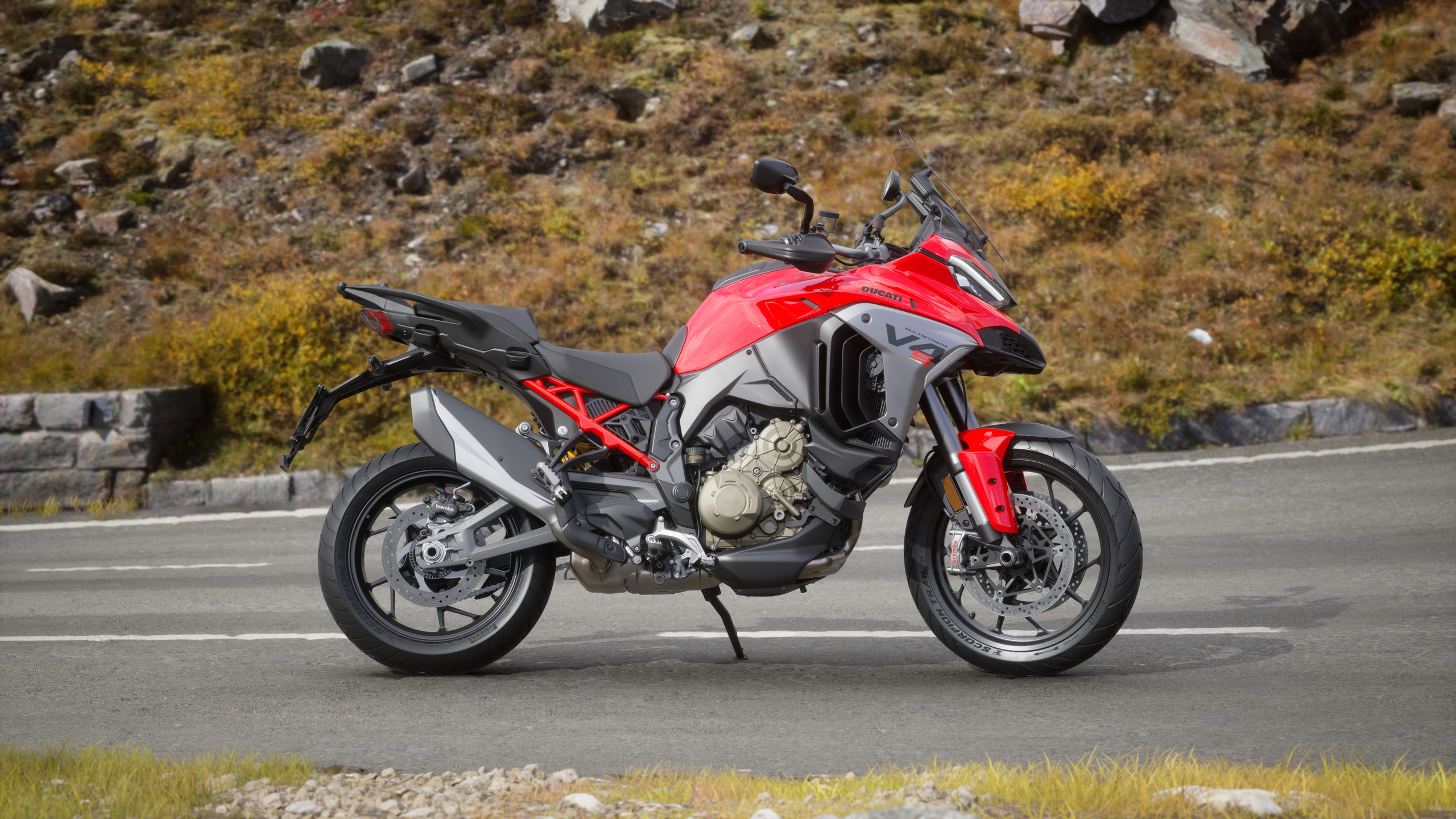 Die individuell konfigurierte Ducati Multistrada V4 in verschiedenen Farben und Ausstattungen zeigt, wie MHPs ECP KI‑generierte Visuals und Gaming‑Technologie mit Produktdaten verknüpft, sodass gewählte Optionen nahtlos bis zur Produktionslinie in Borgo Panigale übernommen werden. <p>Man sieht: Rote Ducati Multistrada V4, Seitenansicht auf dem Seitenständer auf einer Bergstraße; im Hintergrund felsige Böschung mit trockenem Gras und Steinen; sichtbare Details wie Motor, Rahmen, Brembo‑Bremse und Windschutzscheibe, klares Tageslicht.<p>