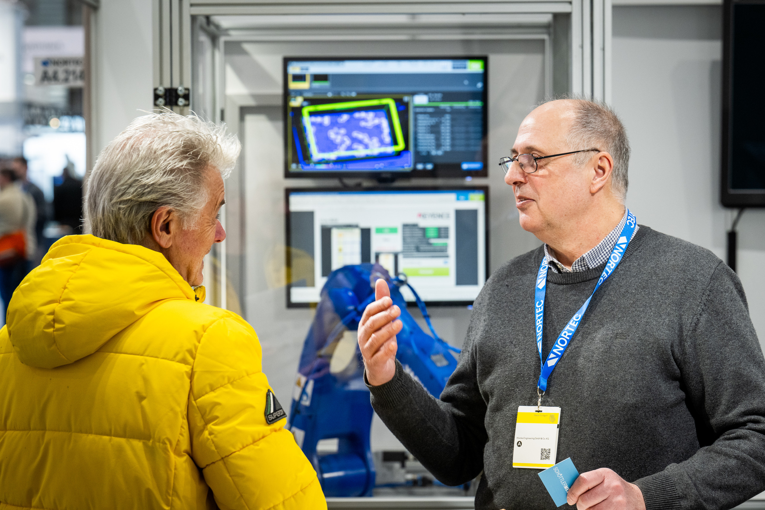 <p>Das Bild zeigt zwei Personen im Gespräch an einem Messestand auf der Nortec 2026. Im Hintergrund sind Bildschirme zu sehen, die vermutlich technische Informationen oder Daten anzeigen. Eine Person trägt eine auffällige gelbe Jacke, während die andere in einem grauen Pulli gekleidet ist. Der Stand scheint sich auf technische Lösungen oder Automatisierung zu konzentrieren.<p>