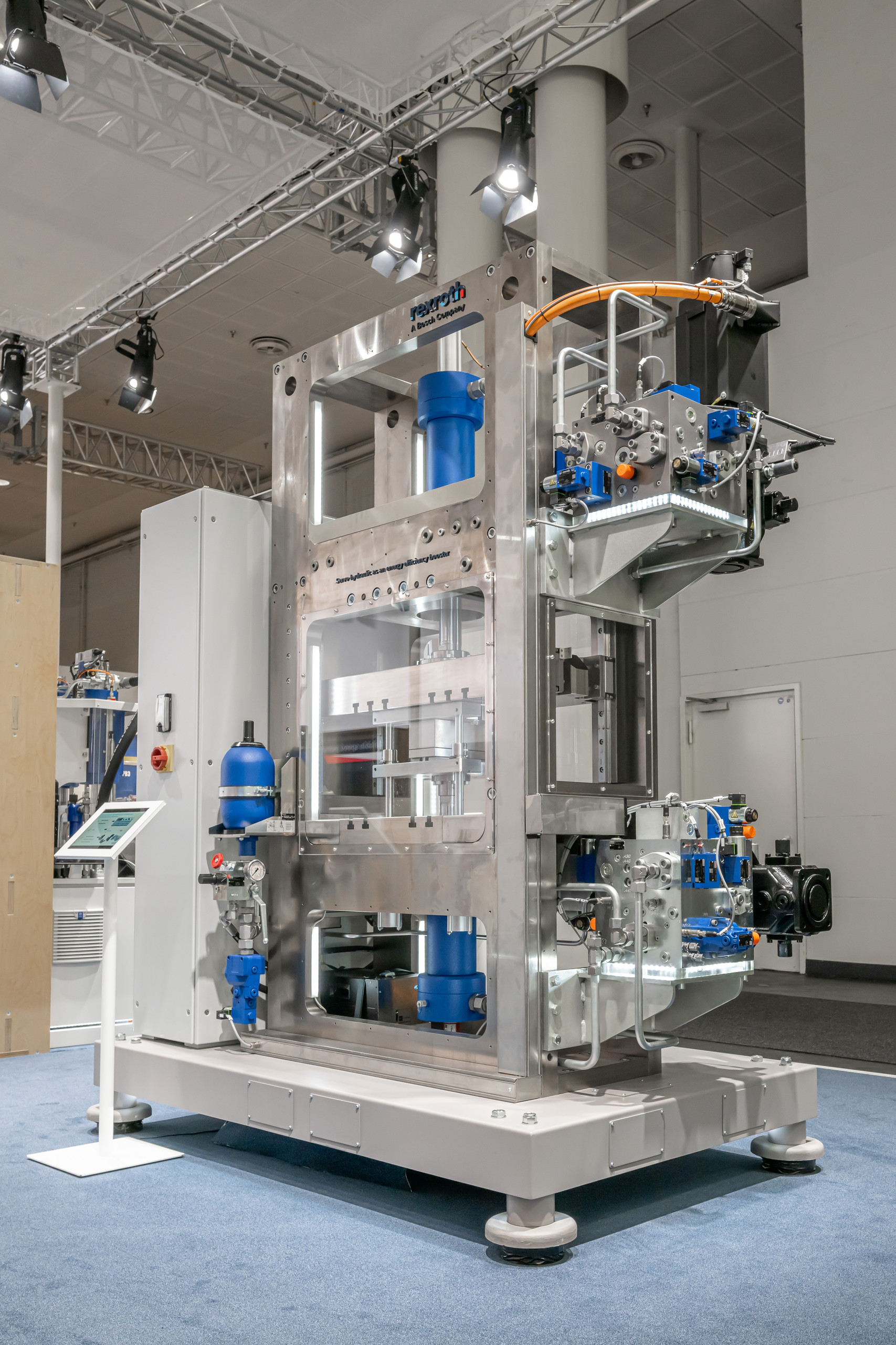 <p>Große Ausstellungsmaschine in einer Messehalle: ein offener Metallrahmen mit Hydraulikzylindern und blauen Komponenten, Steuerkasten, Schläuchen und Verkabelung auf einem Podest; daneben ein Infoständer, darüber Messe‑Scheinwerfer und Traversen.<p>