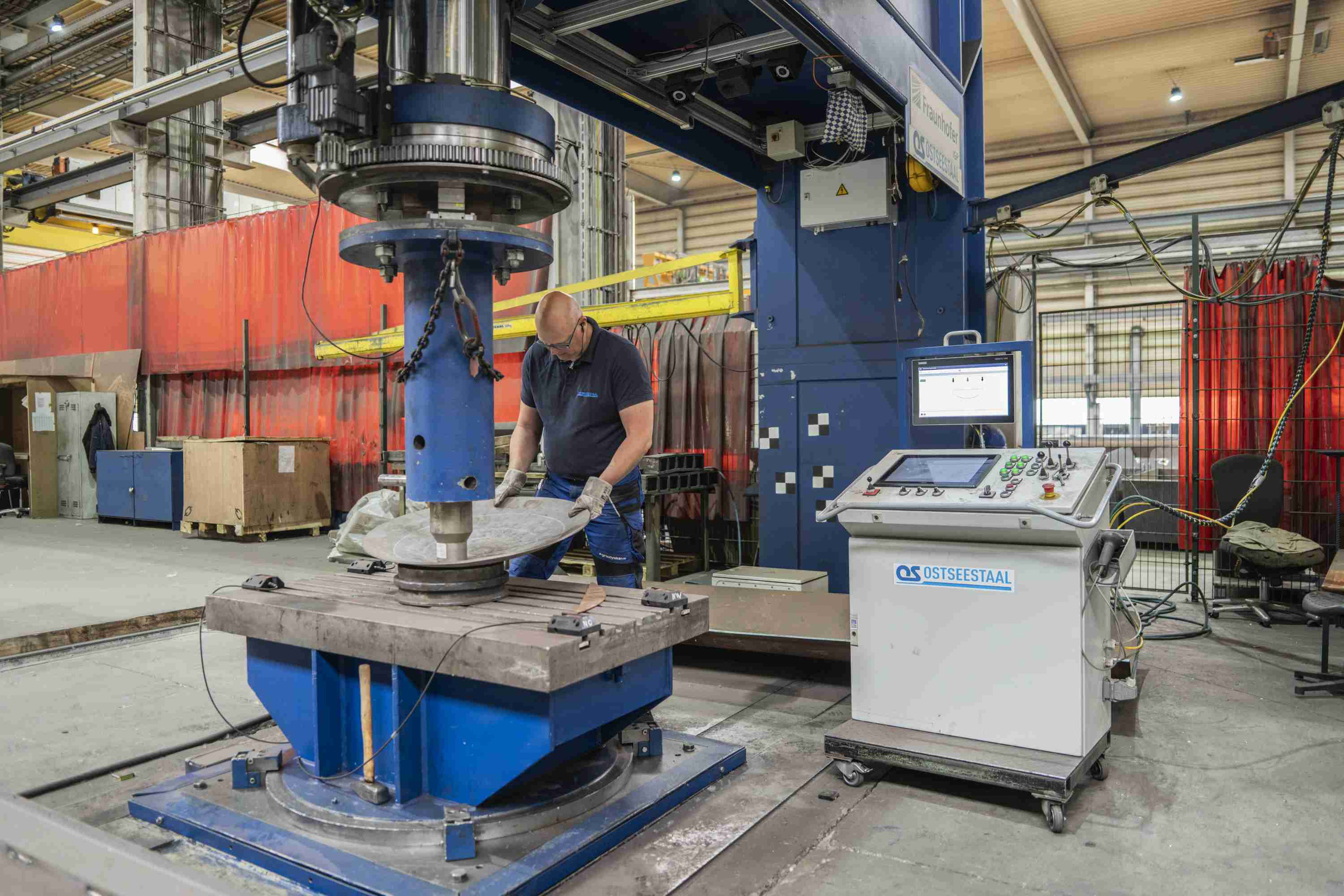 Im Rahmen des Projekts DIKUQ führt ein Werker die Umformung von Stahlblechen durch, um die Effizienz und Qualität in der Fertigung zu verbessern. <p>In dem Bild sieht man eine industrielle Umgebung, in der ein Werker an einer Maschinenanlage arbeitet. Er ist dabei, ein Stahlblech mit einer hydraulischen Presse oder Umformmaschine zu bearbeiten. Auf der rechten Seite gibt es ein Bedienfeld mit einem Bildschirm, das zur Steuerung der Maschine verwendet wird. Die gesamte Einrichtung ist darauf ausgelegt, den Umformungsprozess effizient und präzise zu gestalten.<p>