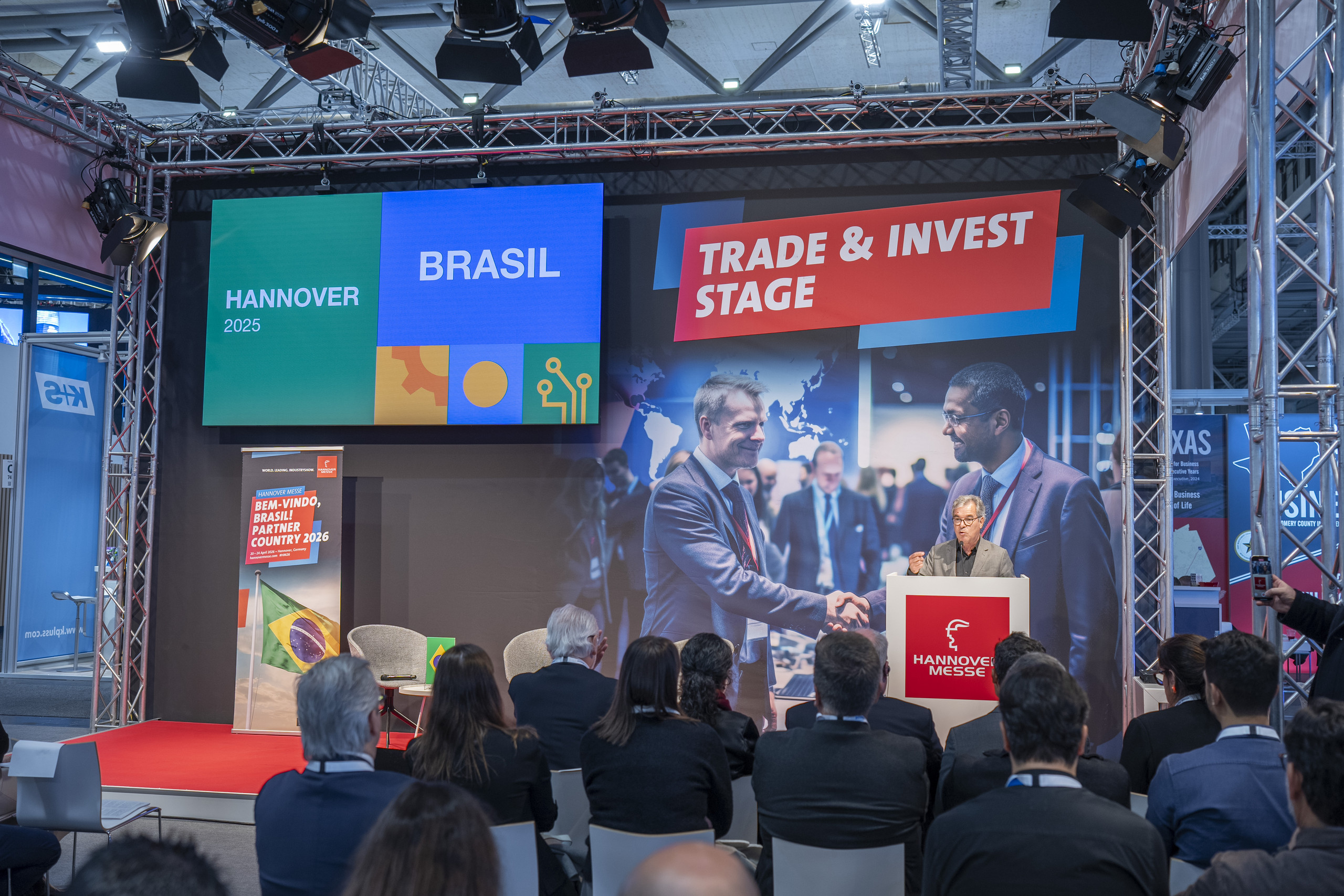 <p>Das Bild zeigt Man eine Bühnen-/Konferenzszene auf der Hannover Messe: Vor einem sitzenden Publikum hält ein Sprecher am Rednerpult (HANNOVER MESSE) einen Vortrag. Hinter ihm hängen große Screens mit der Aufschrift „HANNOVER 2025 – BRASIL“ sowie „TRADE & INVEST STAGE“. Auf dem Bühnenmotiv ist zudem ein großes Bild eines Händedrucks zu sehen – als Symbol für internationale Zusammenarbeit und Investitionen.<p>