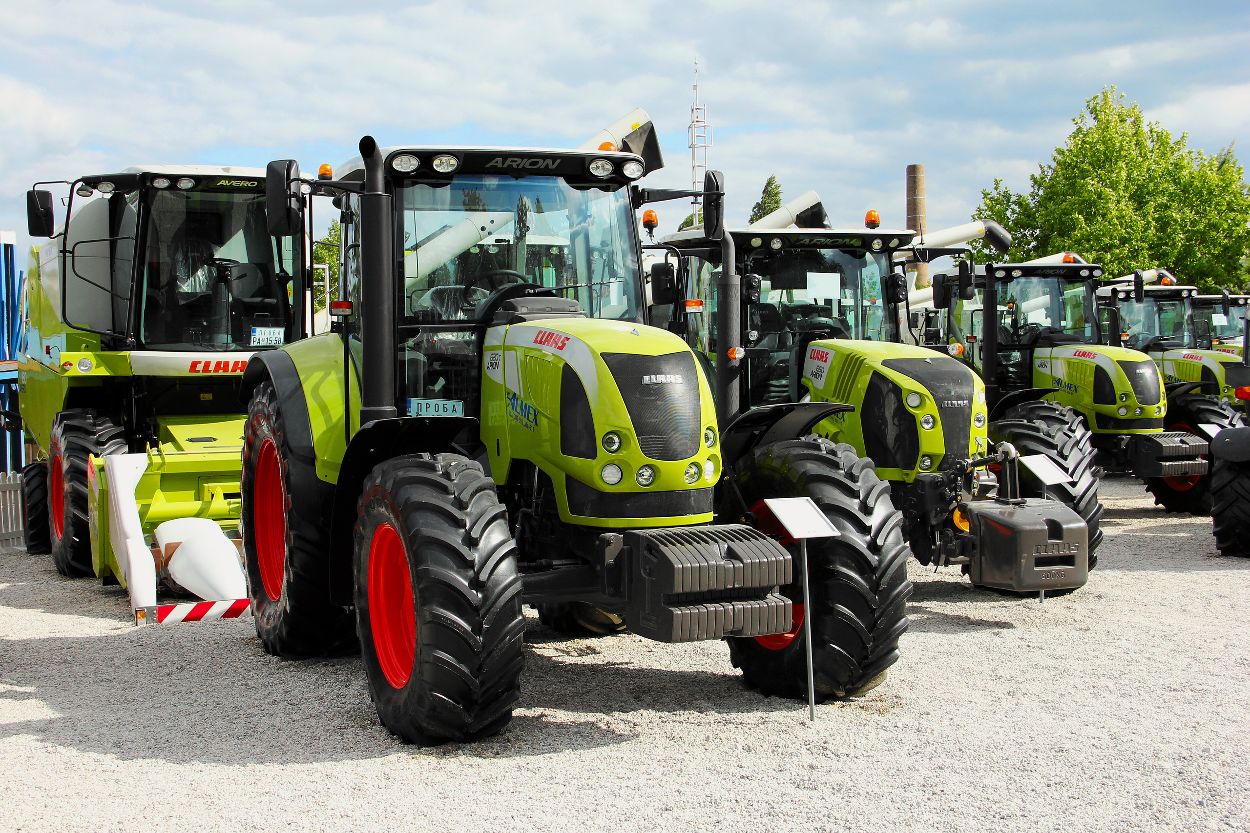 <p>Mehrere moderne, hellgrüne Claas Arion Traktoren mit markanten roten Felgen stehen in einer Reihe auf einem Kiesplatz; im Hintergrund ist weitere Landtechnik zu sehen.<p>
