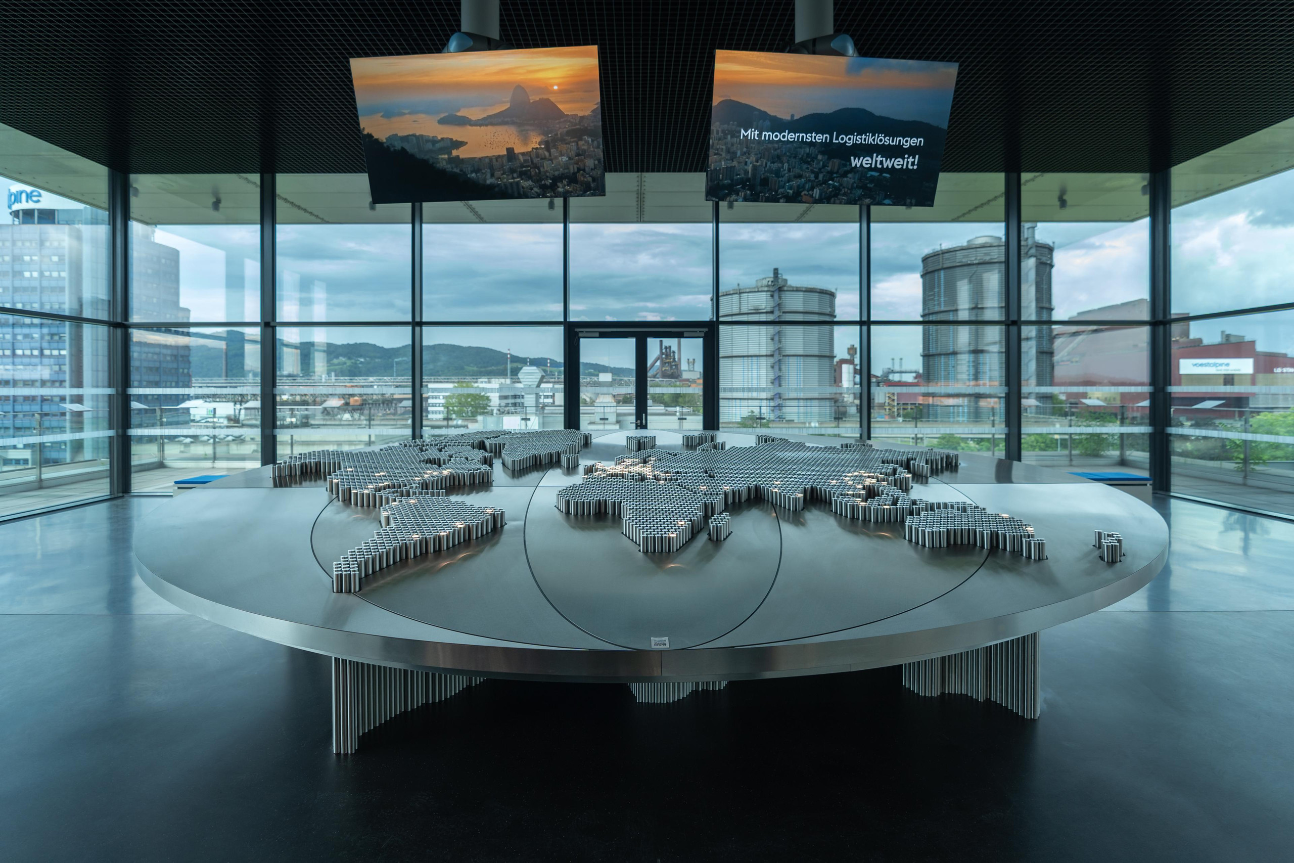 Die stilisierte Weltkarte in der Voestalpine Stahlwelt veranschaulicht die globale Reichweite des Konzerns und bietet Besuchern einen direkten Blick auf das angrenzende Linzer Werksgelände. <p>Auf dem Bild: Ein großer, runder Metalltisch mit einer dreidimensionalen Weltkarte aus unzähligen vertikalen Metallstiften vor einer Panorama-Glasfront, die den Blick auf die Industrieanlagen der Voestalpine freigibt.<p>