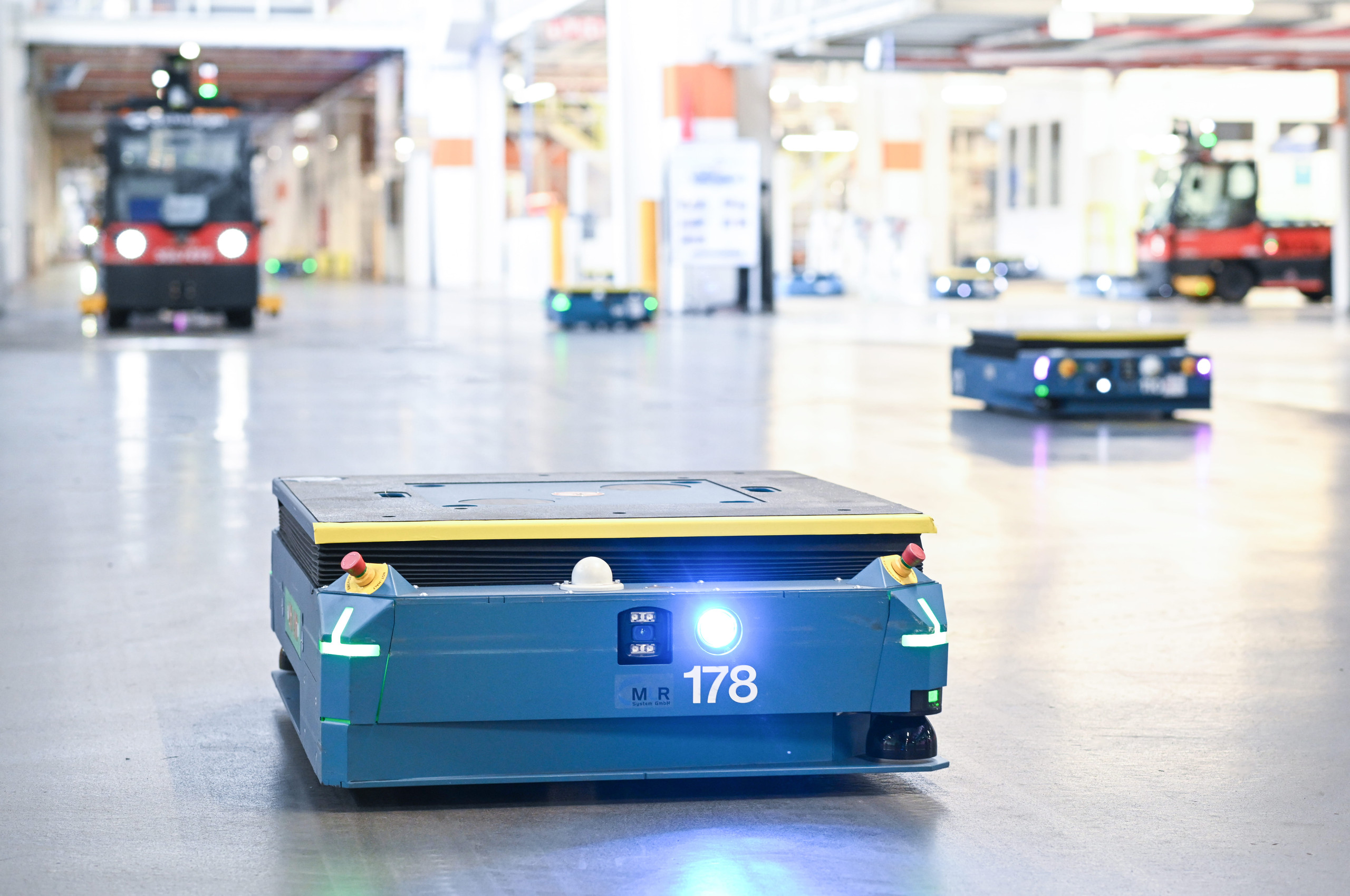 <p>Blick in eine Industriehalle mit verschiedenen Transportmitteln: Im Vordergrund ein blauer autonomer Transportroboter (AMR), dahinter weitere Roboter und ein roter Gabelstapler. Die Szene zeigt eine typische heterogene Flottenlandschaft, in der unterschiedliche Systeme auf gemeinsamen Fahrwegen agieren.<p>