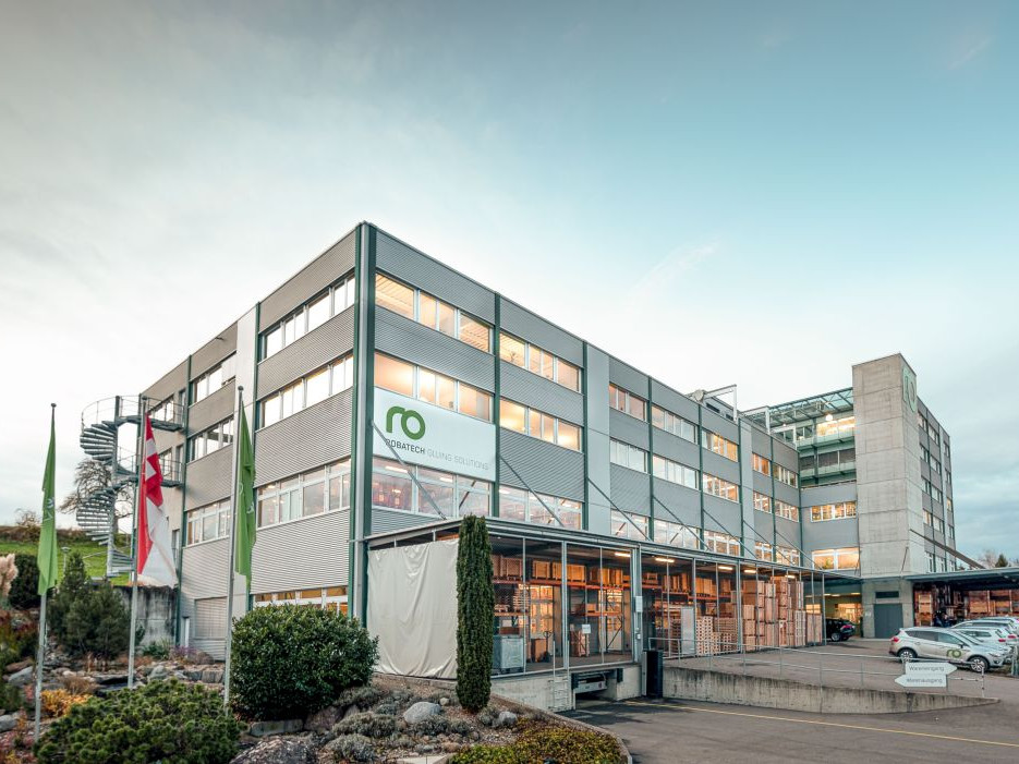  Das Bild zeigt die Fassade eines modernen Geschäftshauses mit mehreren Etagen, das von großen Fenstern geprägt ist. Das Gebäude besitzt einen glatten, silbernen Metalllook und trägt das Logo von "Robatech" an einer prominenten Stelle. Vor dem Eingang sind stilvolle Pflanzen und Flaggen zu sehen. Eine auffällige spiralförmige Treppe führt zu einem oberen Bereich. Im Vordergrund parken mehrere Autos, und der Himmel ist in sanften Farben gehalten, was eine einladende Atmosphäre schafft.