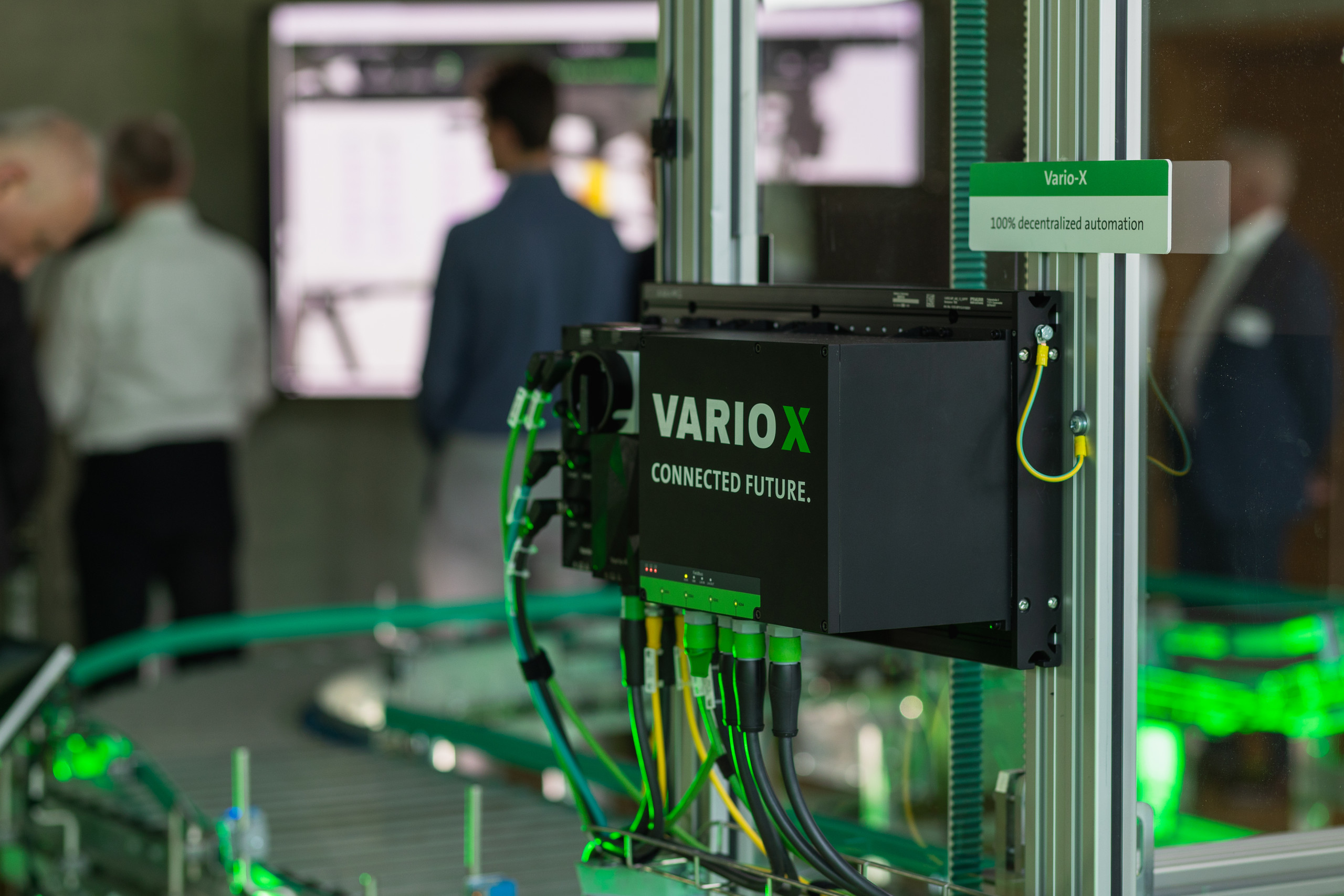 <p>Man sieht: Nahaufnahme der dezentralen Automatisierungsplattform Vario-X von Murrelektronik. Das schwarze Steuerungsgehäuse mit der Aufschrift „VARIO-X CONNECTED FUTURE“ ist an einer silbernen Profilschiene montiert. An der Unterseite sind mehrere schwarze und grüne Kabel über kompakte MQ15-Steckverbinder angeschlossen. Im Hintergrund ist eine industrielle Anwendung mit Förderband und Personen in einer Werkshalle unscharf erkennbar.<p>