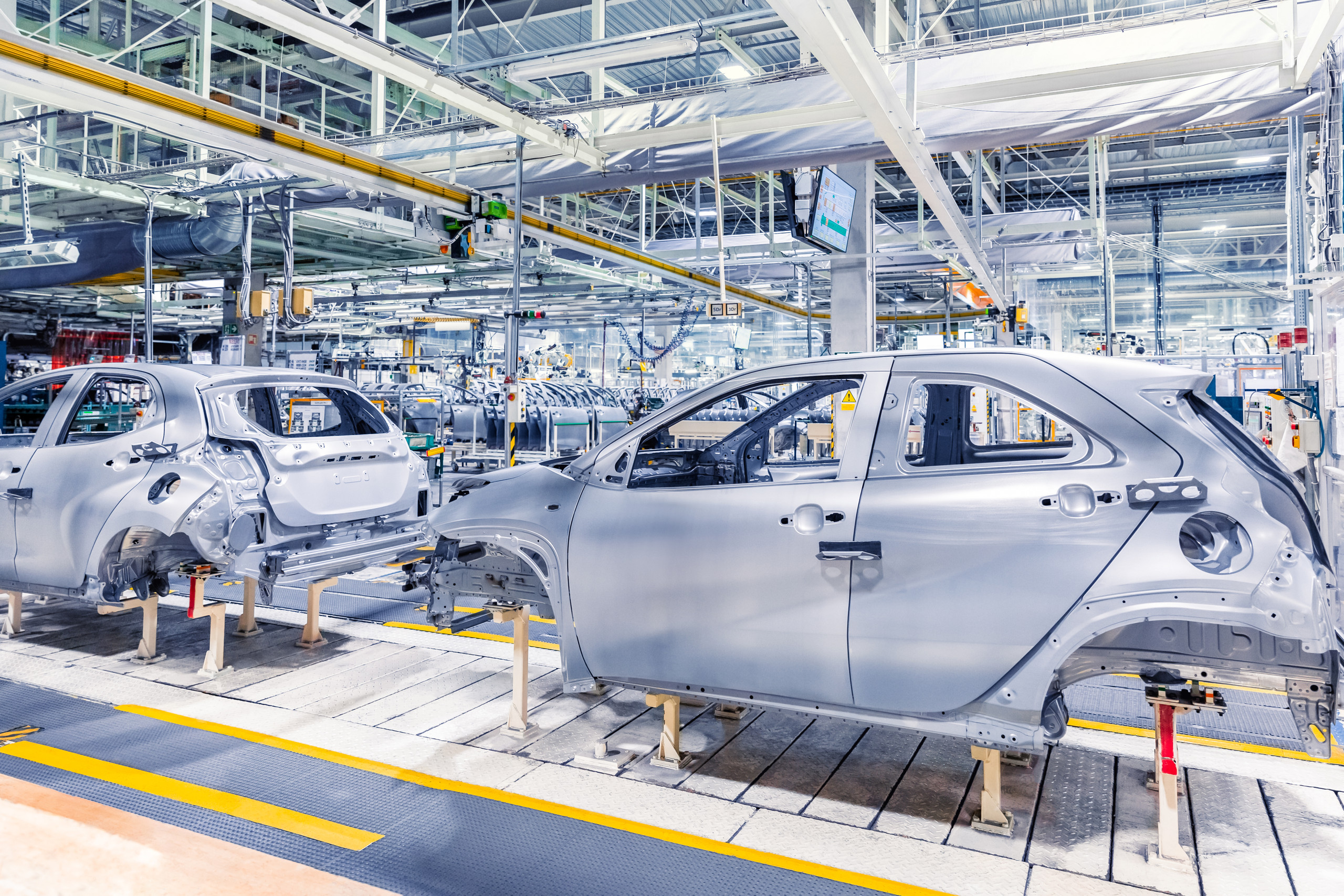 <p>Das Bild zeigt eine Automobilproduktionslinie mit unvollständigen Fahrzeugkarosserien, die auf Ständern platziert sind. Der Raum ist hell erleuchtet und hat eine moderne, industriell gestaltete Umgebung. Über den Karosserien sind verschiedene Förder- und Automatisierungssysteme sichtbar, was auf einen hochgradig automatisierten Fertigungsprozess hinweist. Die Szene vermittelt ein Gefühl von Effizienz und Technologie in der Automobilproduktion.<p>