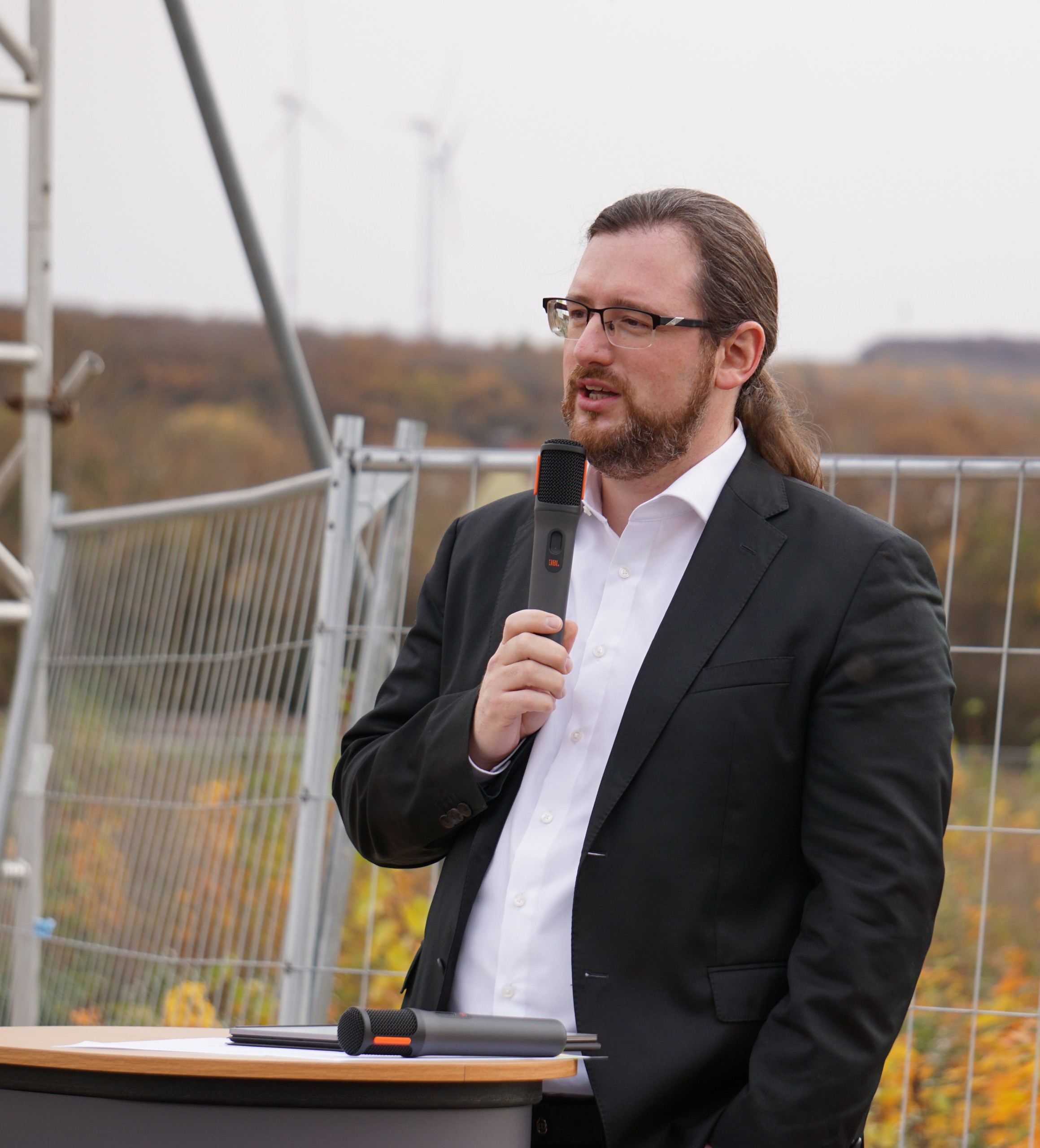 Mann mittleren Alters mit Anzug und zum Zopf zusammengebundenen braunen langen Haaren spricht auf einer Baustelle zu den Gästen