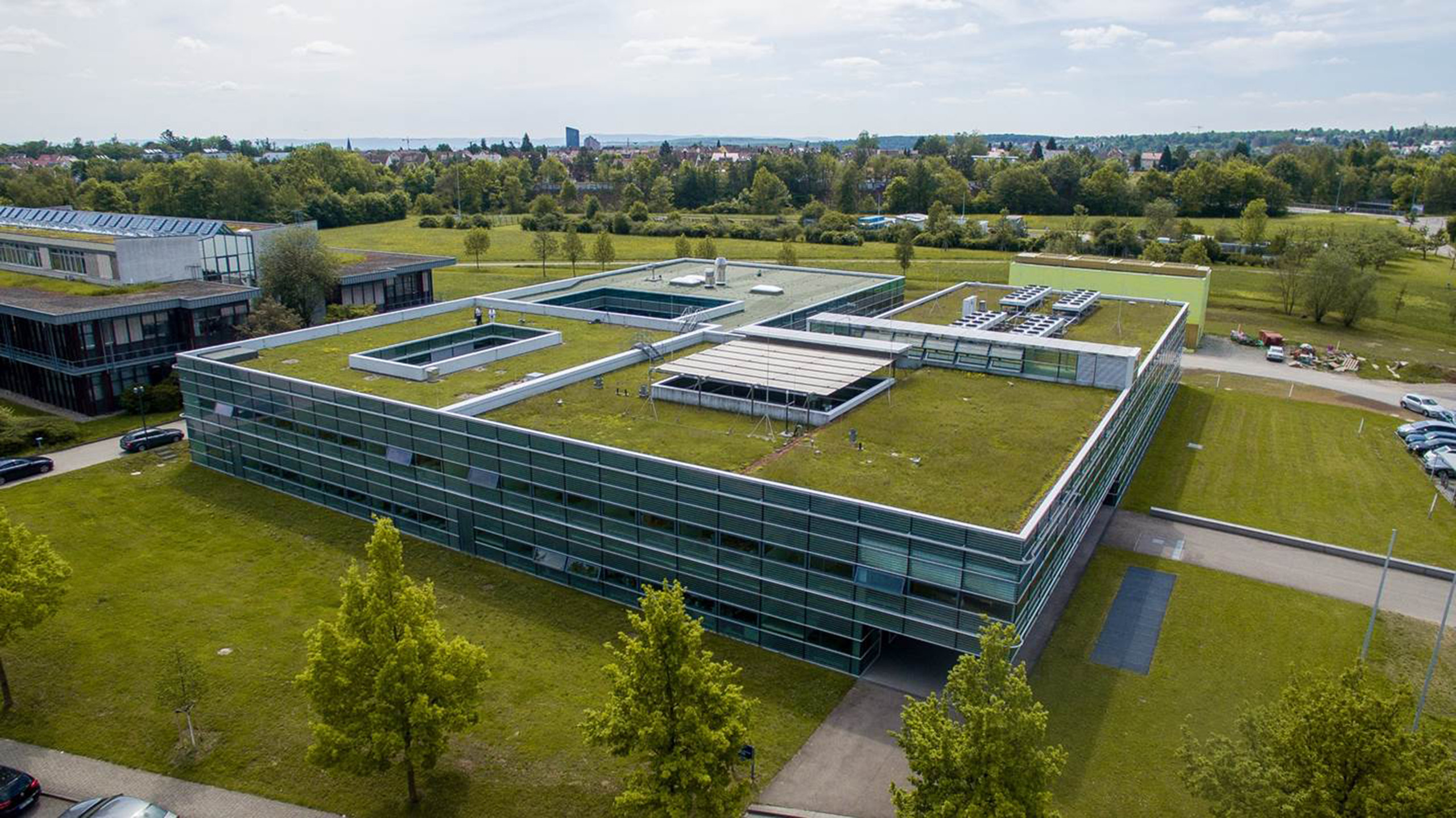 <p>Man sieht: Vogelperspektive auf ein modernes, flaches Büro- oder Forschungsgebäude mit einer gläsernen Außenfassade. Das Dach ist komplett mit grünem Rasen und Pflanzen bewachsen und verfügt über mehrere rechteckige Lichtschächte bzw. Innenhöfe. Das Gebäude steht auf einer gepflegten Rasenfläche mit Bäumen; im Hintergrund ist eine bewaldete Landschaft unter hellem Himmel zu sehen.<p>