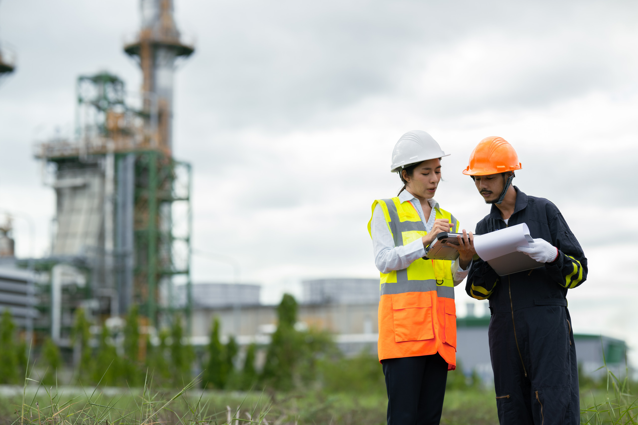 <p>Man sieht: Zwei Personen stehen im Vordergrund einer Industrieanlage; eine trägt eine weiße Schutzhelm und eine gelb‑orange Warnweste, die andere einen orangefarbenen Schutzhelm und Arbeitskleidung. Gemeinsam betrachten sie ein Tablet und technische Pläne, eine handgehaltene Seite wird von einer Person mit Handschuh gehalten. Im unscharfen Hintergrund sind Türme, Rohrleitungen und Tanks einer Raffinerie/Industrieräumlichkeit sichtbar, davor Gras und ein bewölkter Himmel.<p>