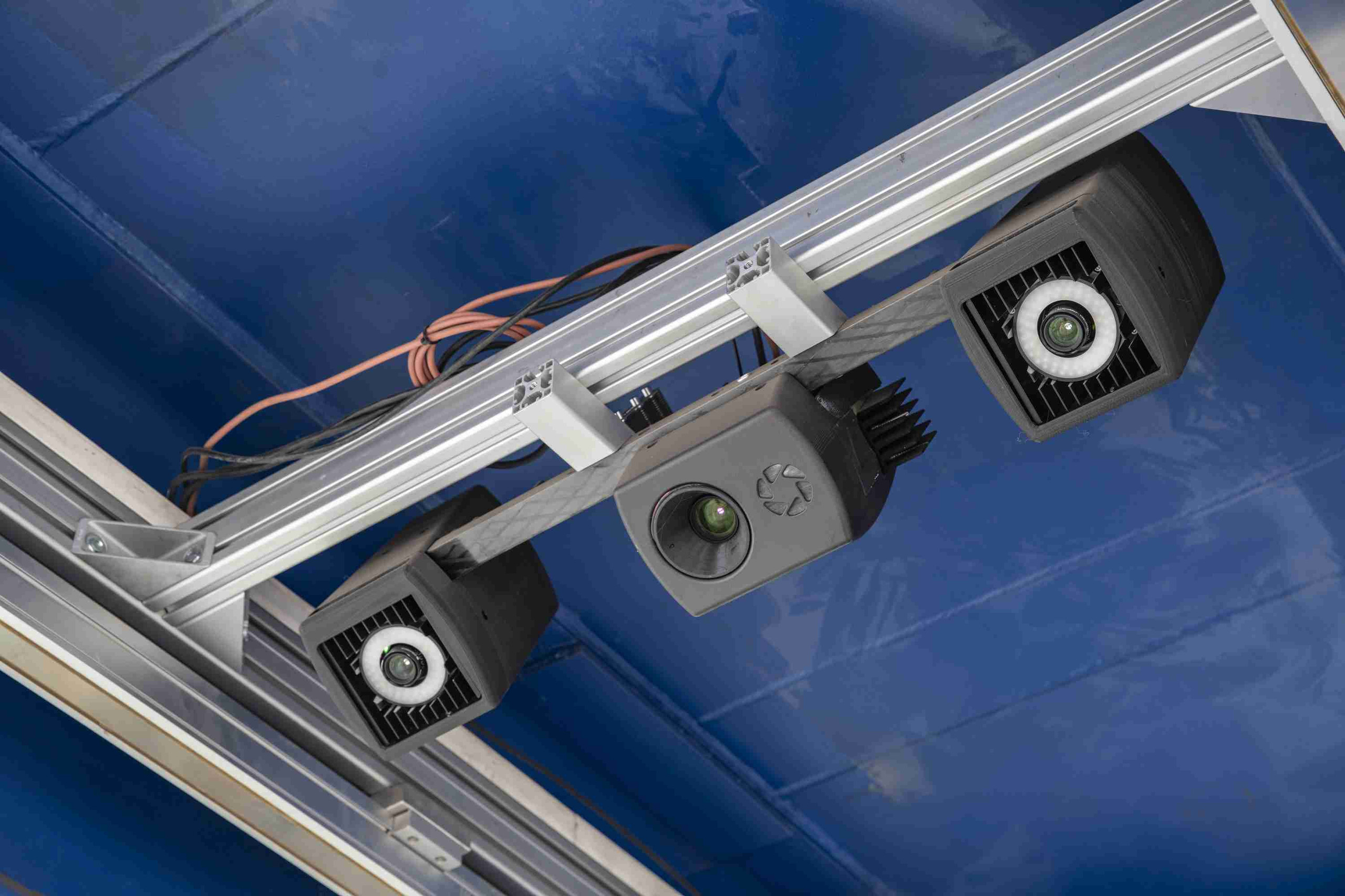 Das Sensornetzwerk besteht aus zueinander synchronisierten Kameras und je einem Projektor zur Musterprojektion für die digitale 3D-Erfassung. <p>Auf dem Bild sieht man eine technische Vorrichtung mit mehreren Kameras oder Sensorsystemen, die an einer Schiene montiert sind. Diese Geräte scheinen für die Überwachung oder Messung in einer industriellen Umgebung konzipiert zu sein. Der Hintergrund ist blau, eventuell ein Teil der Werkstattausstattung. Die Kameras sind wahrscheinlich Teil eines Inline-3D-Messsystems zur digitalen Erfassung von Objekten oder Werkstücken.<p>