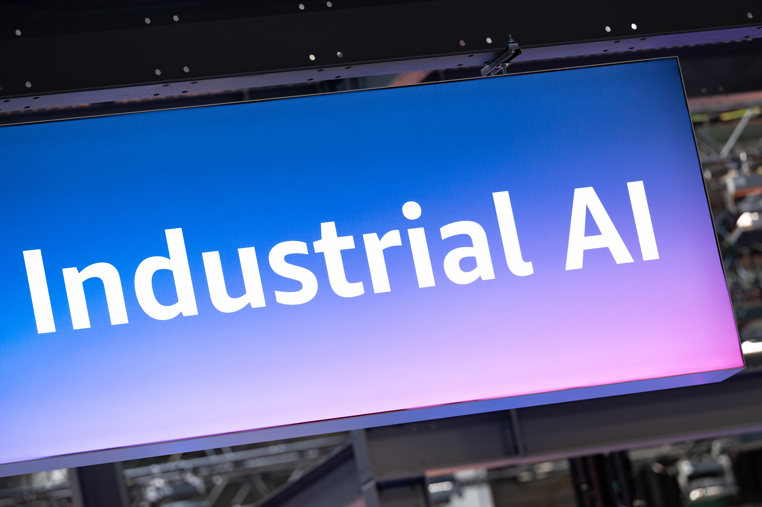 <p>Auf dem Bild sieht man ein beleuchtetes Banner mit dem Schriftzug „Industrial AI“ in großen, weißen Buchstaben. Der Hintergrund des Banners ist in einem sanften Farbverlauf von Blau zu Lila gehalten. Das Banner scheint Teil einer Messe oder Ausstellung zu sein, die sich mit Themen der künstlichen Intelligenz im industriellen Sektor beschäftigt. Im unteren Teil des Bildes sind Elemente einer Messeinfrastruktur sichtbar, was darauf hindeutet, dass es sich um eine Veranstaltung handelt.<p>