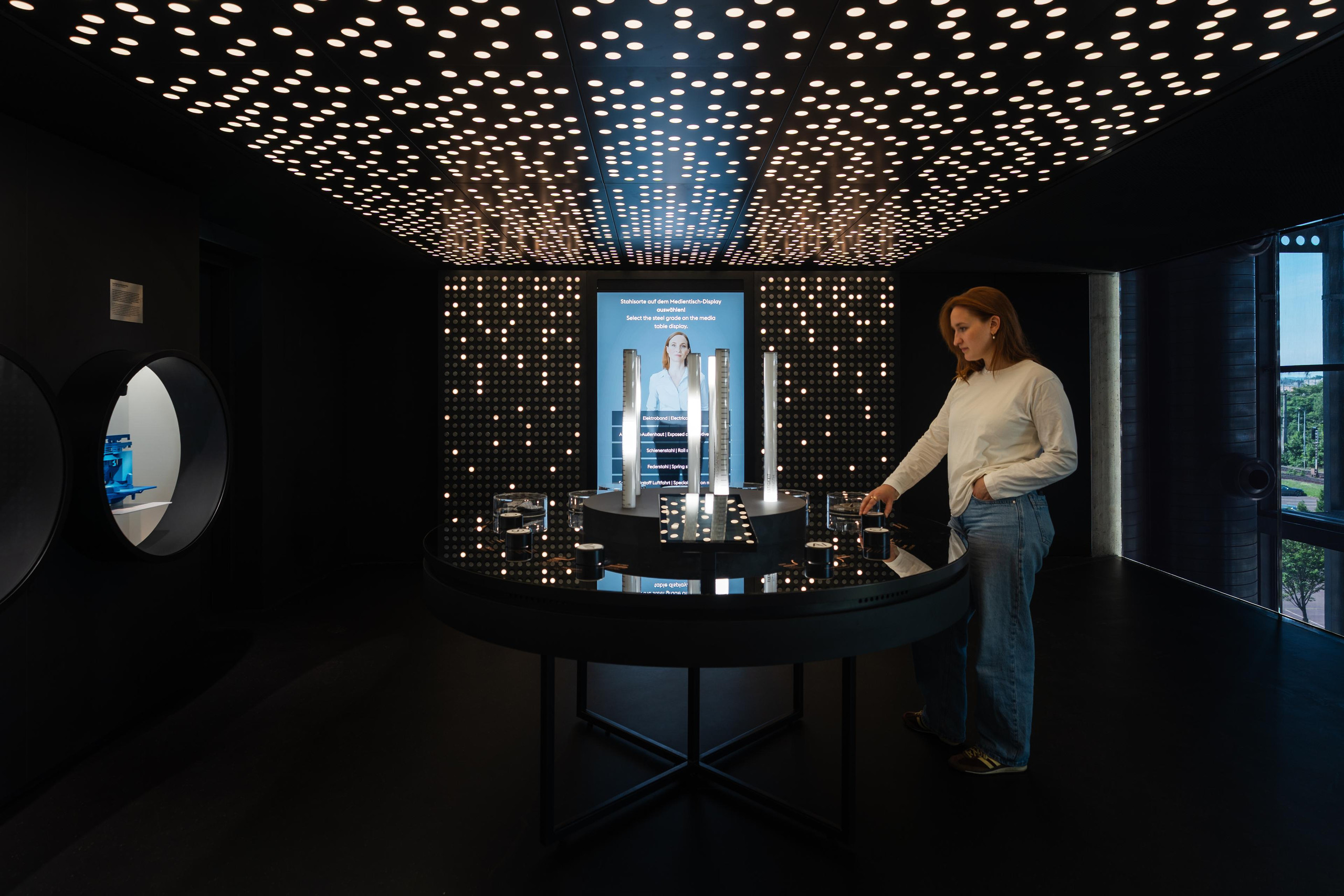 Der interaktive Legierungstisch in der voestalpine Stahlwelt ermöglicht es Besuchern, die komplexen Prozesse der Stahlerzeugung und Materialzusammensetzung spielerisch zu entdecken. <p>Auf dem Bild: Eine Frau interagiert mit einem runden, schwarzen Medientisch in einem dunkel gestalteten Ausstellungsraum der Voestalpine Stahlwelt, während die Decke über ihr von unzähligen kreisförmigen Lichtpunkten erleuchtet wird.<p>
