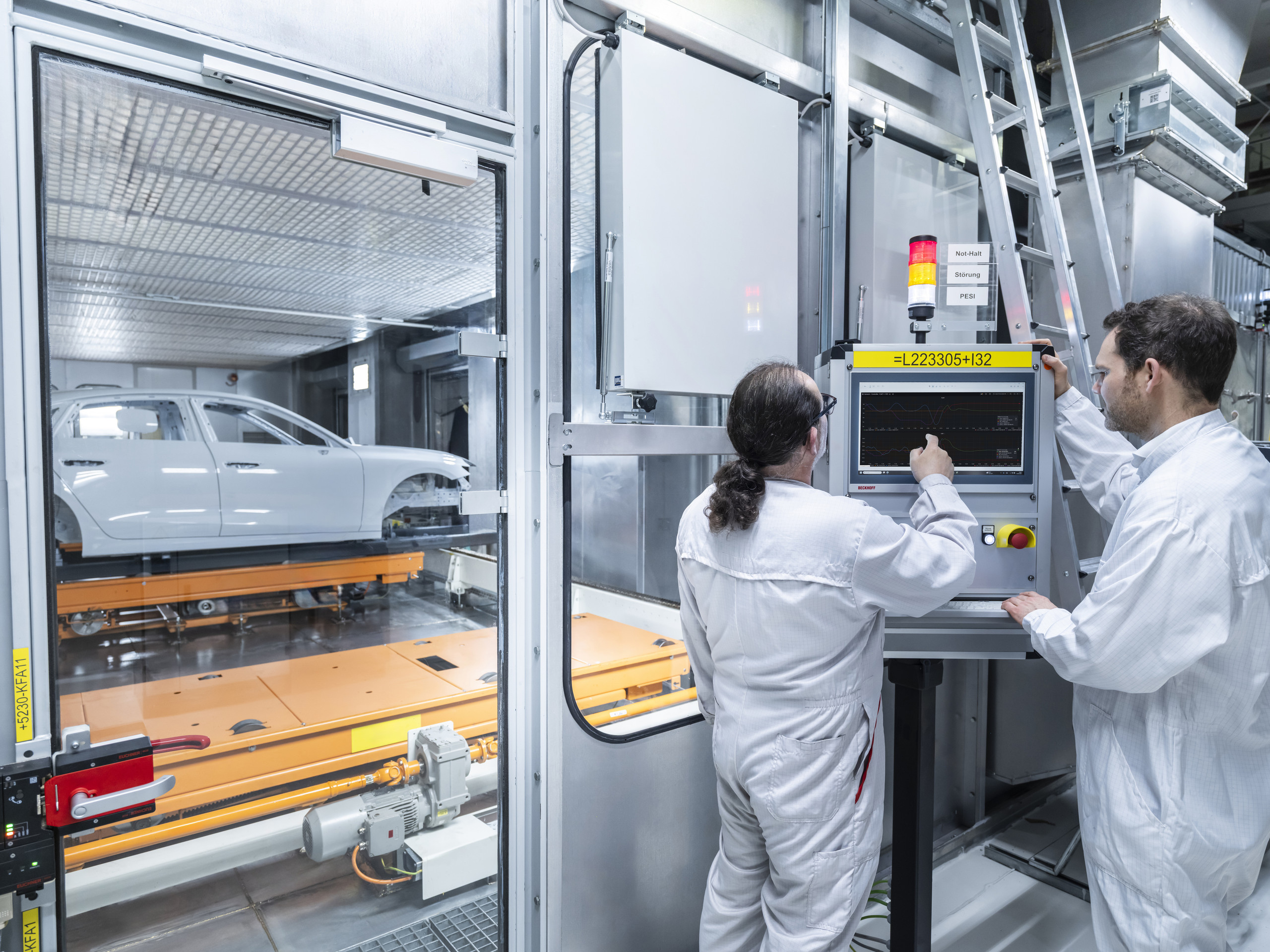 Audi Neckarsulm KI-Steuerung Trockner zwei Beschäftigte an einem Industrie-PC vor einer Scheibe, hinter der Scheibe wird eine Rohkarosse in den Trockner befördert