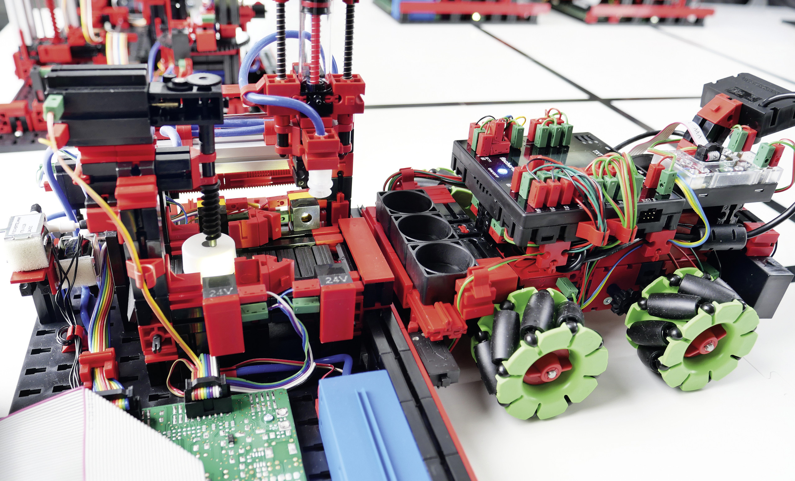 <p>Man sieht: Detailansicht der Agile Production Simulation: Ein mobiles Transportfahrzeug mit Mecanum-Rädern dockt an einer automatisierten Bearbeitungsstation mit komplexer Verkabelung und Sensortechnik an.<p>