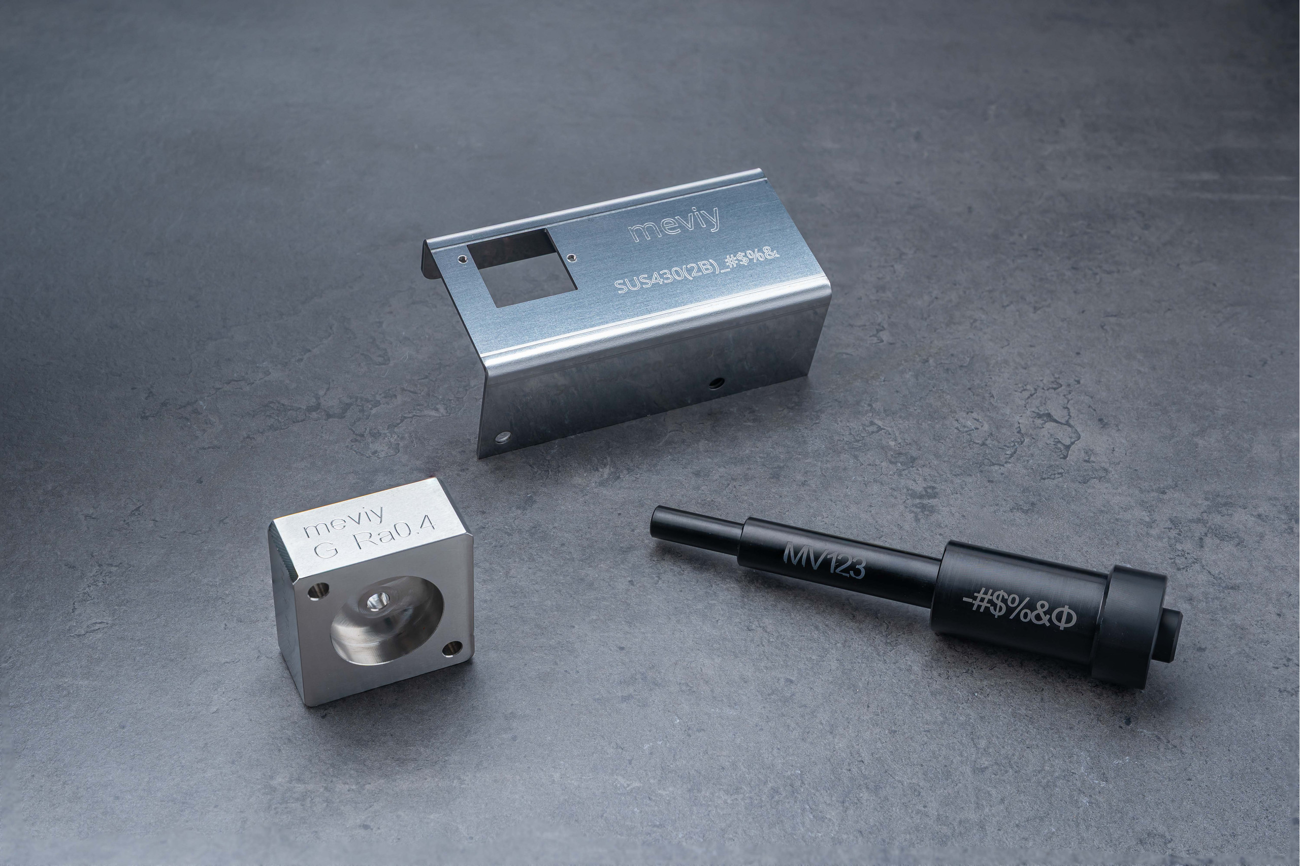 <p>Auf dem Bild sind drei verschiedene metallische Bauteile zu sehen, die vermutlich für industrielle Anwendungen konzipiert sind:  Oben (rechtes Bauteil): Ein längliches, geformtes Metallteil mit einer offenen Fläche und mehreren gravierten Zeichen, das möglicherweise eine Art Gehäuse oder Halterung darstellt.  Mitte (quadratisches Bauteil): Ein kompaktes, quadratisches Metallstück mit einer tiefen Vertiefung auf einer Seite und gravierten Beschriftungen. Es könnte sich um einen Halter oder eine Verbindungskomponente handeln.  Unten (schwarzes Bauteil): Ein zylindrisches, schwarzes Metallteil, das wie ein Stift oder eine Buchse aussieht und ebenfalls angedeutete Gravuren aufweist. Es könnte für Befestigungs- oder Montagesysteme verwendet werden.  Alle Teile befinden sich auf einem grauen, strukturierten Untergrund, der das Metall gut zur Geltung bringt.<p>