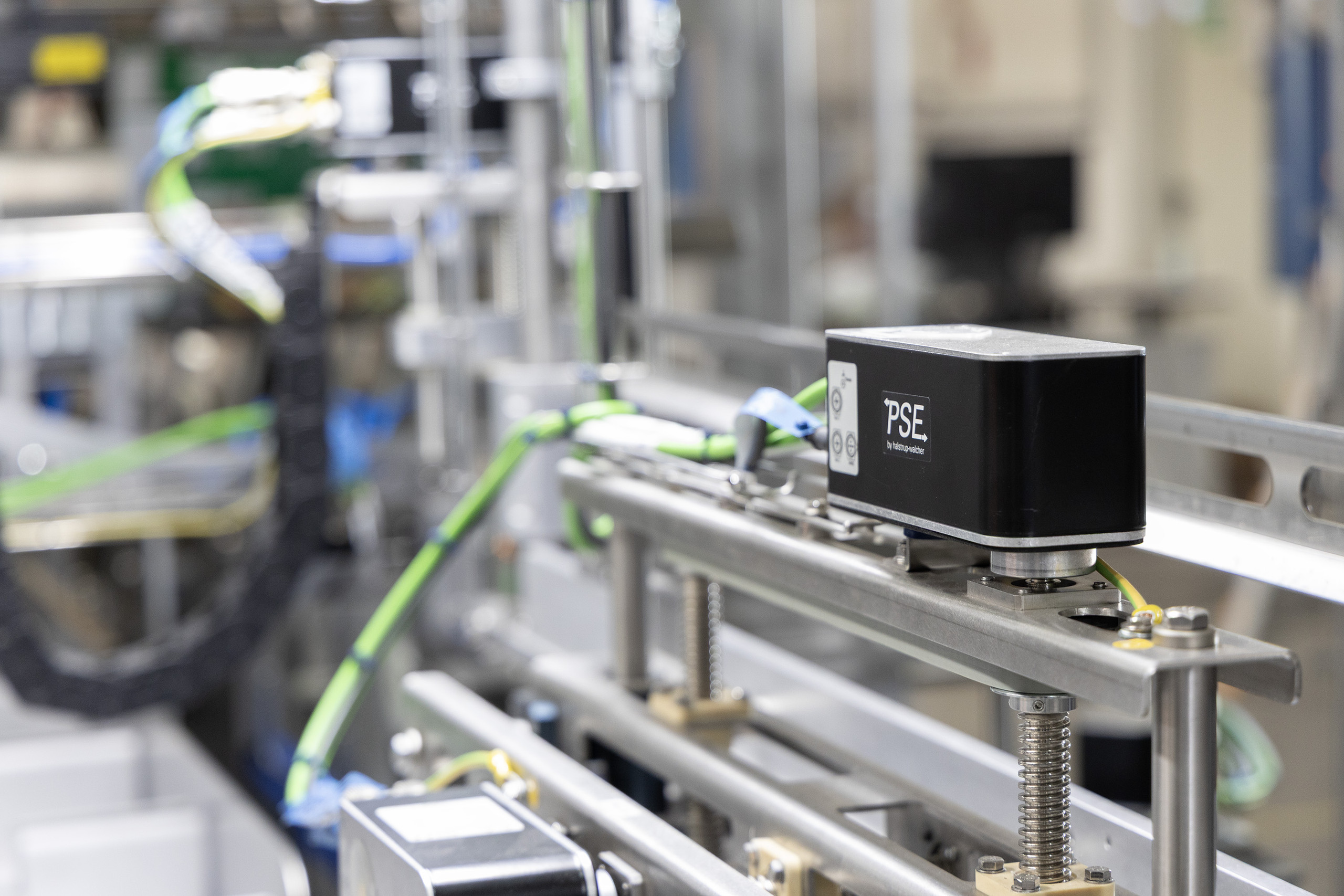 <p>Man sieht ein mechanisches Bauteil, das offenbar Teil einer automatisierten Maschine ist. Im Vordergrund ist ein schwarzes Gerät mit der Aufschrift "PSE" zu sehen, das wahrscheinlich ein Stellantrieb oder Sensor ist. Es ist auf einer metallischen Schiene montiert, umgeben von verschiedenen Kabeln und Komponenten. Dies deutet auf eine präzise Steuerung innerhalb eines industriellen Prozesses hin.<p>