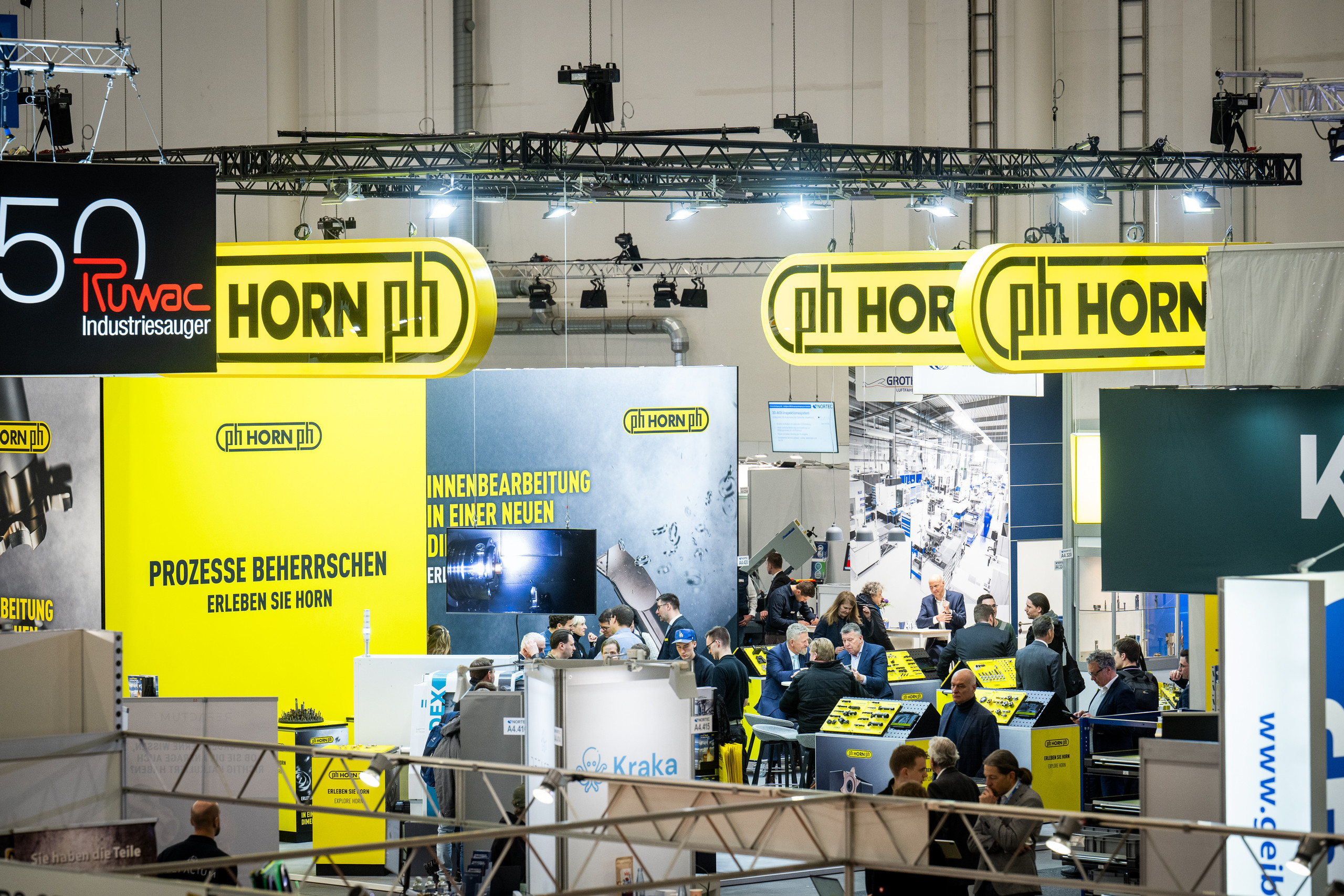 <p>der Abbildung sind verschiedene Fertigungswerkzeuge und -komponenten ausgestellt. Die Präsentation erfolgt auf roten Ablagen in einem Messestand, wobei folgende Elemente zu sehen sind:  Verschiedene Werkzeuge: Dazu gehören Präzisionswerkzeuge mit unterschiedlichen Formen und Größen, die für verschiedene Fertigungsprozesse geeignet sind. Montage- und Frästeile: Mechanische Komponenten, die für den Maschinenbau oder die Automation verwendet werden. Gespäche im Hintergrund: Fachbesucher und Aussteller, die sich über die Produkte austauschen, wodurch die interaktive Atmosphäre der Messe verdeutlicht wird. Die gesamte Szene vermittelt einen Eindruck von Innovation und technischem Austausch in der Industrie.<p>