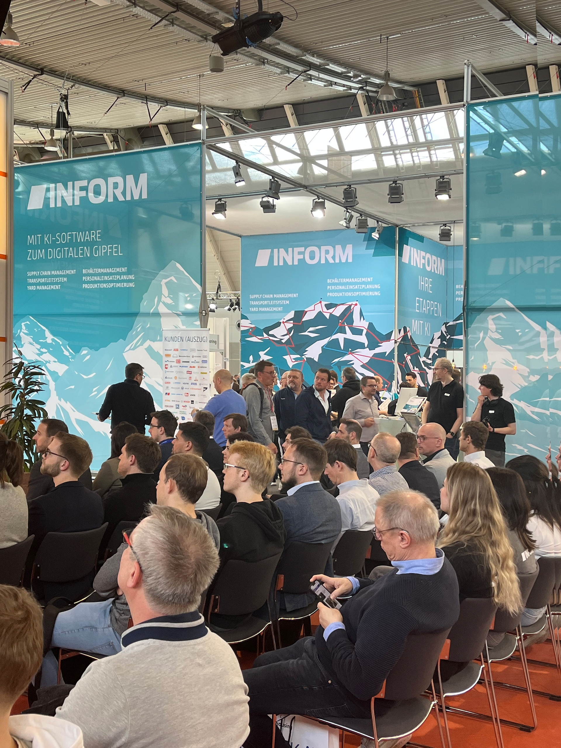 <p>Das Bild zeigt einen Messestand der Firma Inform auf einer Veranstaltung. Im Vordergrund sind zahlreiche Besucher zu sehen, die auf Stühlen sitzen und dem Geschehen am Stand folgen. Im Hintergrund sind die Banner des Unternehmens mit Slogans wie „Mit KI-Software zum digitalen Gipfel“ und Angeboten wie Supply Chain Management und Transportoptimierung zu sehen. Der Bereich wirkt lebhaft, und es scheint eine Präsentation oder Diskussion am Stand stattzufinden. Die Umgebung ist hell und modern, mit einem roten Teppichboden.<p>