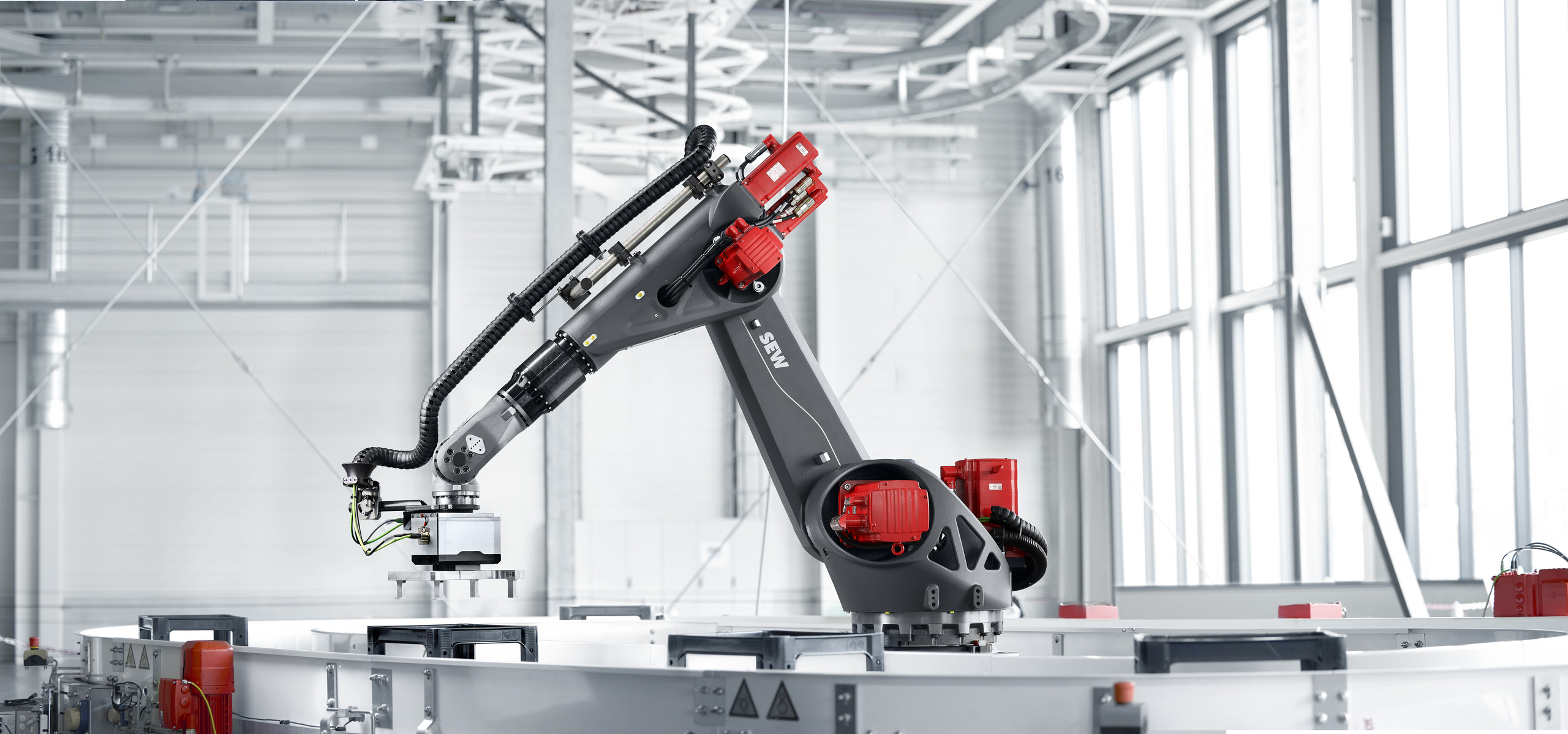 Knickarmroboter aus dem Baukasten von SEW-EURODRIVE lassen sich präzise und zuverlässig in bestehende Produktionslinien integrieren. <p>Grau‑roter Knickarmroboter (SEW‑Logo sichtbar) hebt mit einem Vakuumgreifer ein Bauteil über einem runden Förder‑/Palettiertisch; im Hintergrund große Fenster und Hallendecke mit Leitungen.<p>