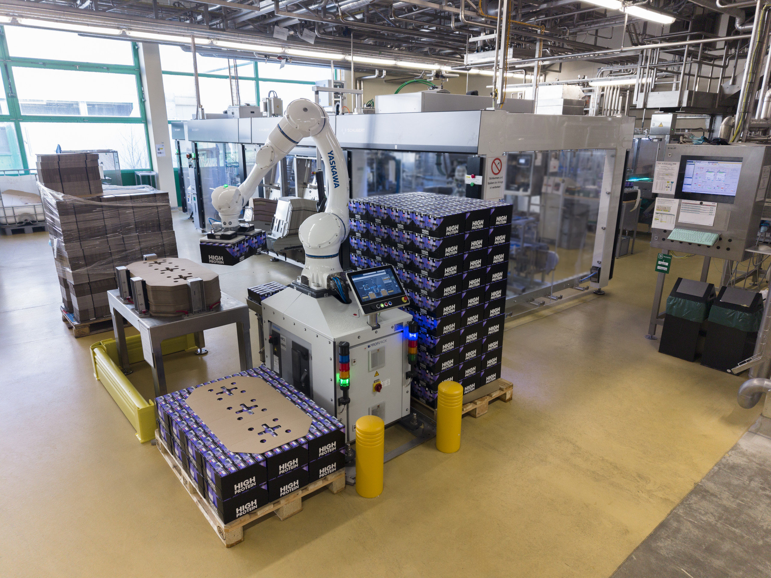 <p>Weitwinkelaufnahme einer kompakten Palettierstation in einer Industriehalle. Ein weißer Yaskawa-Cobot ist zentral auf einer Steuereinheit von Profipack montiert und stapelt schwarze Joghurt-Trays auf Euro-Paletten. Die Anlage ist frei zugänglich, benötigt keine Schutzzäune und zeigt im Vordergrund einen Stapel mit Karton-Zwischenlagen sowie gelbe Anfahrschutz-Poller.<p>