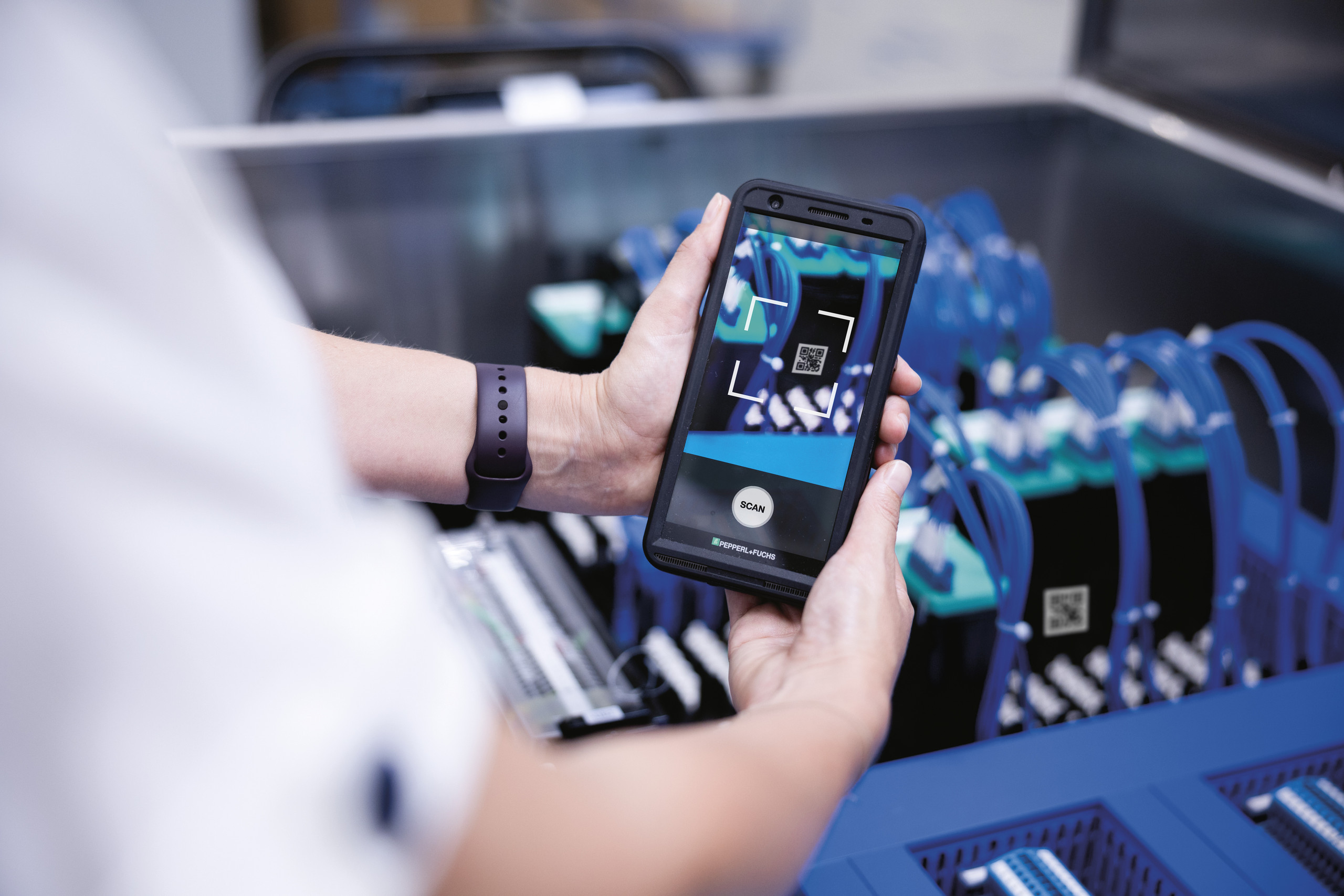 <p>Man sieht: Hände halten ein Smartphone, das einen QR‑Code auf einer elektrischen Baugruppe mit blauen Kabeln scannt; unscharfe Produktionsumgebung im Hintergrund.<p>
