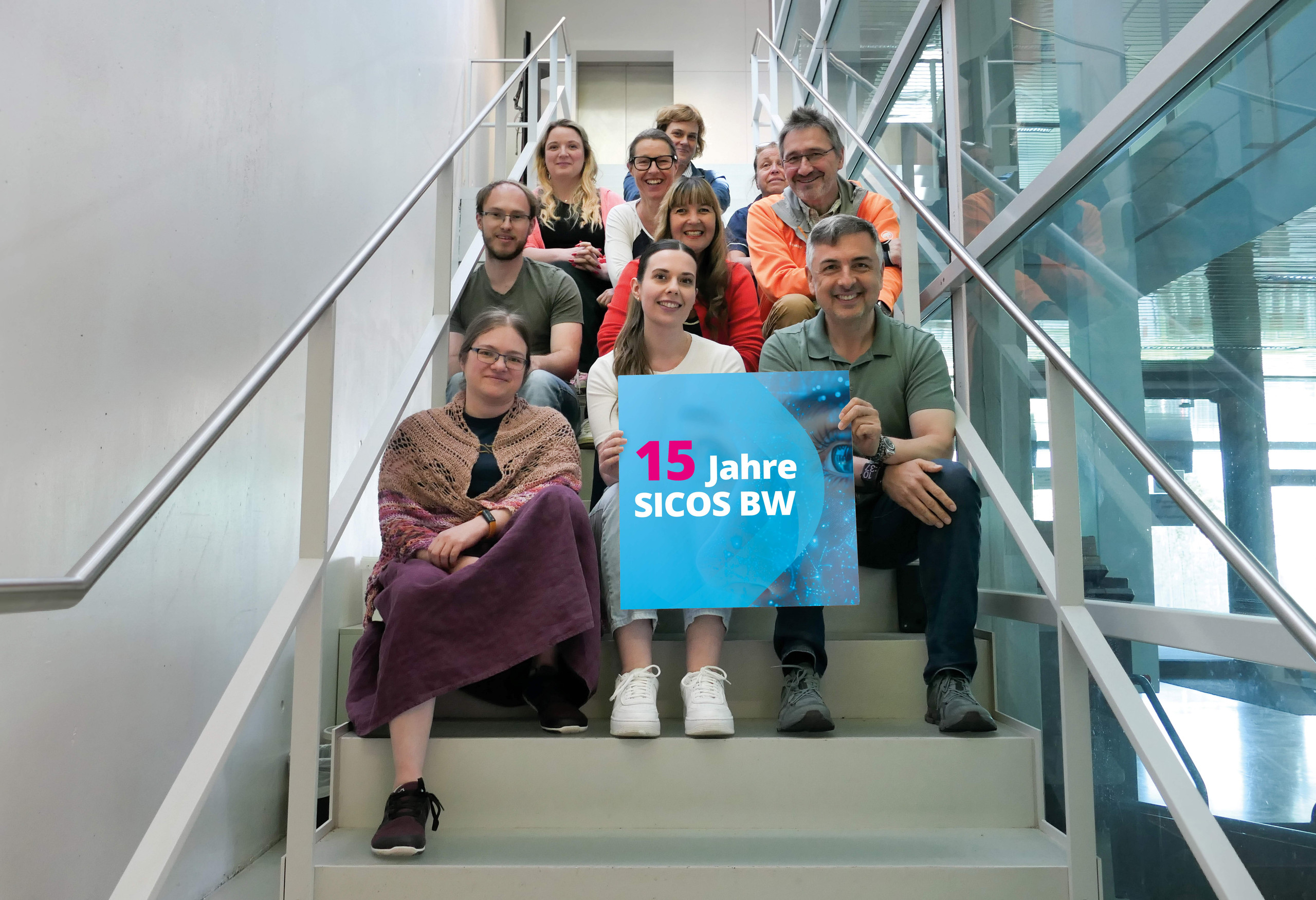 <p>Man sieht: Gruppenfoto des Sicos BW Teams auf einer Treppe. In der Bildmitte halten eine Frau und ein Mann gemeinsam ein quadratisches blaues Schild mit der Aufschrift „15 Jahre SICOS BW“. Das Team lächelt in die Kamera und symbolisiert die persönliche Beratung rund um digitale Innovationen.<p>