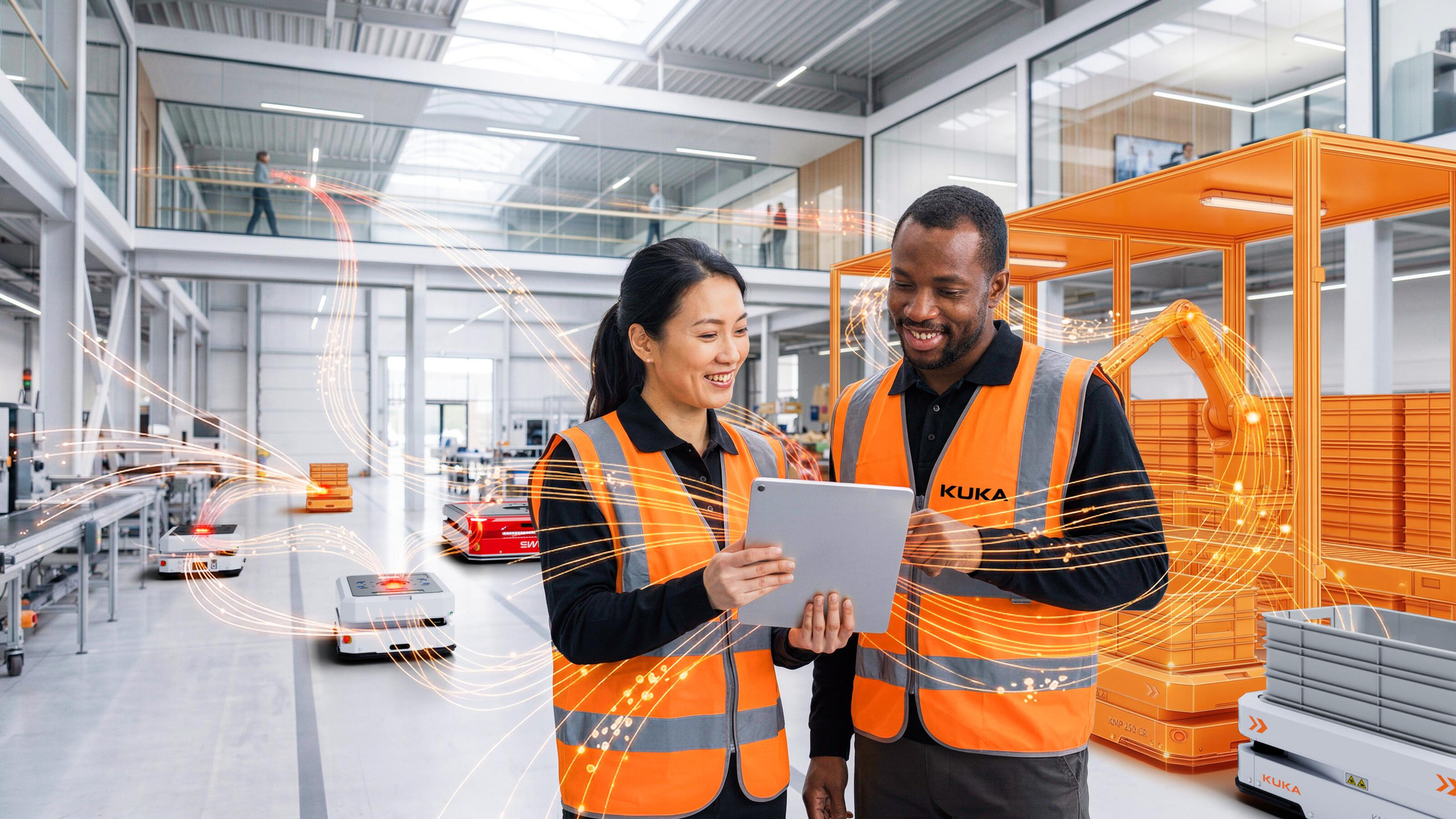 <p>Man sieht: Innenaufnahme einer hellen, modernen Fabrikhalle: im Vordergrund stehen zwei Mitarbeitende in orangefarbenen Schutzwesten und betrachten gemeinsam ein Tablet. Dahinter sind mehrere autonome mobile Roboter (AMR) auf dem Hallenboden, Fördertechnik, Behälter und ein in Kontur gehaltener Roboterarm zu erkennen. Über das Bild verlaufen orangefarbene Lichtspuren, die Datenaustausch und KI‑gestützte Vernetzung zwischen Mensch, Maschine und Materialfluss symbolisieren. Die Szene zeigt die Zusammenarbeit von Personal und autonomen Systemen in einer digital vernetzten Produktionsumgebung.<p>