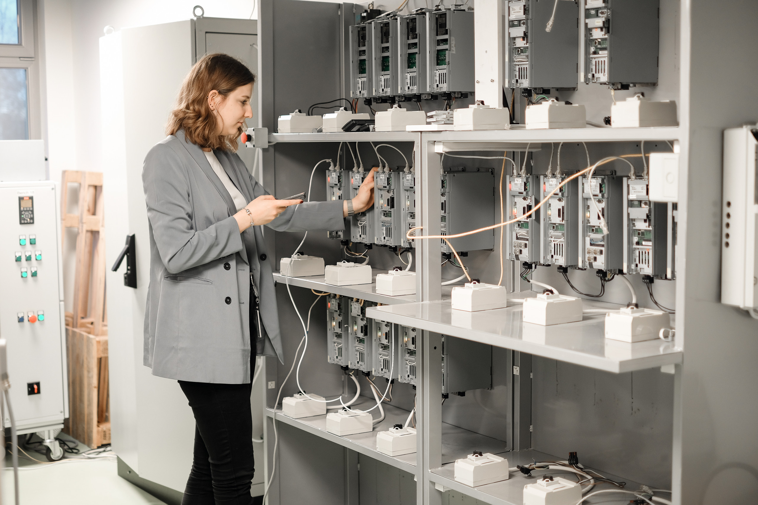 Bestehende Produkte lassen sich durch gezielte Softwareanpassungen sicher und CRA-konform weiterentwickeln – ganz ohne Eingriffe in die Hardware. <p>Eine Frau in grauem Blazer steht an einem Prüfgestell und hält ein Smartphone, während sie mit der anderen Hand an eines von mehreren geöffneten Elektronik‑/Steuergeräten an einem Regalgehäuse arbeitet; zahlreiche Kabel und weiße Anschlusskästchen sind sichtbar.<p>