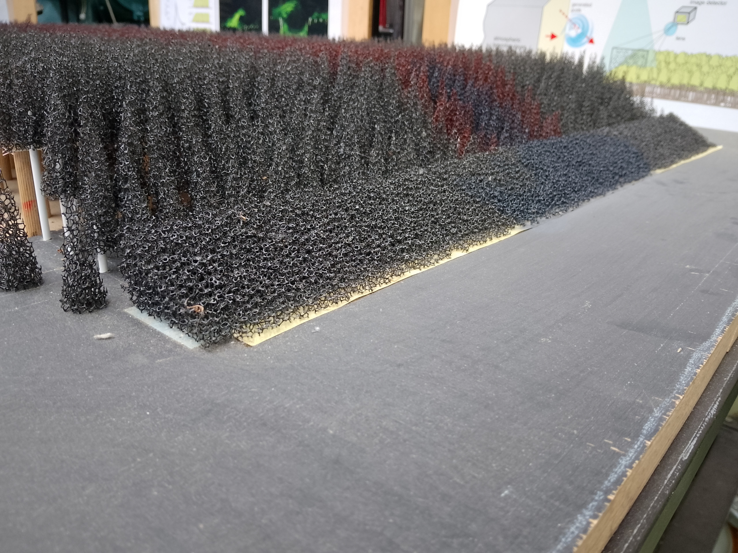 <p>Auf dem Bild ist ein Modell oder eine experimentelle Anordnung zu sehen, das aus zahlreichen dünnen, spiralförmigen Strukturen besteht, die schwarz gefärbt sind. Diese Strukturen scheinen eine Art von Umgebungsmodell zu repräsentieren, möglicherweise im Zusammenhang mit strömungsmechanischen Untersuchungen, etwa zur Simulation von Vegetation oder anderen Hindernissen für die Luftströmung. Der Untergrund scheint eine Holzplatte zu sein, und im Hintergrund sind möglicherweise weitere Materialien oder Informationsgrafiken sichtbar. Die Anordnung könnte in einem Forschungs- oder Bildungslabor des Karlsruher Instituts für Technologie (KIT) verwendet werden.<p>