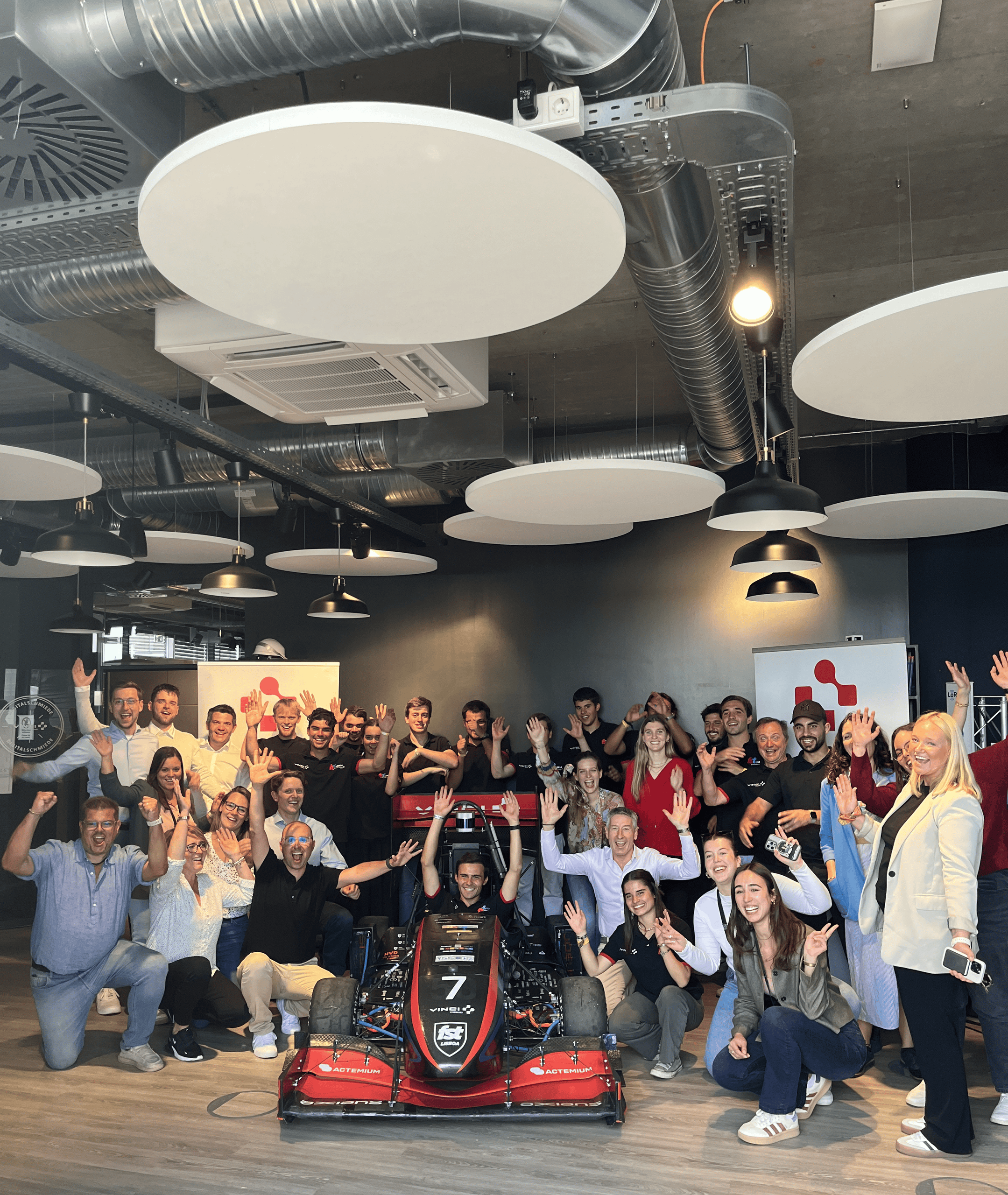<p>Man sieht: Gruppenfoto mit rund 30 Personen in einem modern‑industriellen Veranstaltungsraum jubeln und winken in die Kamera, während in der Mitte ein rotes Formel‑Rennauto (Nr. 7) mit VINCI/Actemium‑Logos steht; Deckenpaneele, Hängeleuchten und Banner im Hintergrund.<p>