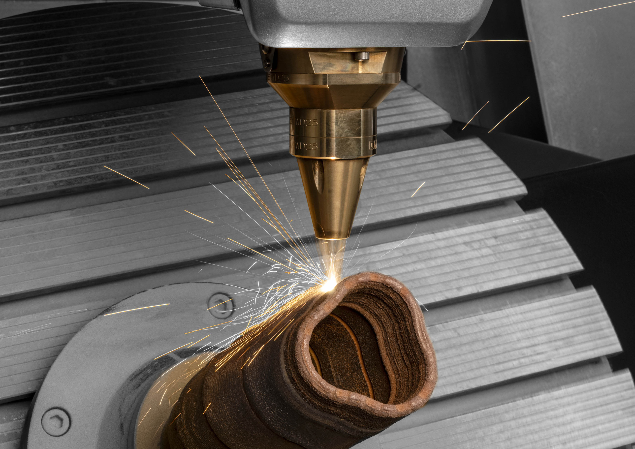 <p>Man sieht: Additive Fertigung mit DMG MORI: Eine LASERTEC DED hybrid Maschine beim Laserauftragschweißen eines komplexen Metallbauteils mit sichtbarem Funkenflug.<p>