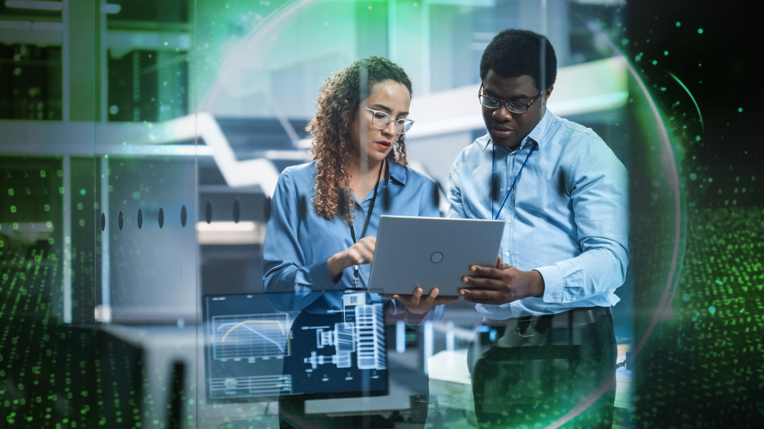 <p>Zwei Personen in einem modernen Technikbüro betrachten gemeinsam ein Laptop. Grüne, halbtransparente Digital‑Overlays und Diagramme liegen über der Szene.<p>