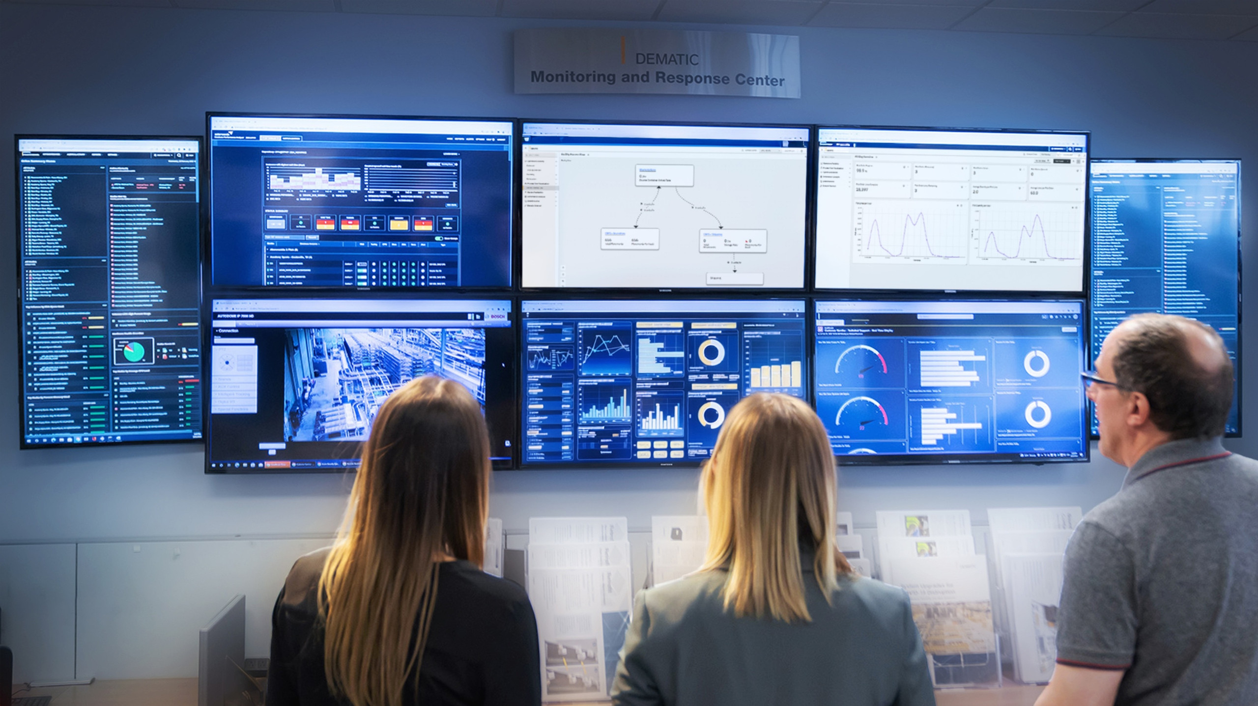 <p>Auf dem Bild: Drei Mitarbeitende im Dematic Monitoring and Response Center blicken auf eine große Monitorwand mit verschiedenen Daten-Dashboards und Echtzeit-Analysen.<p>