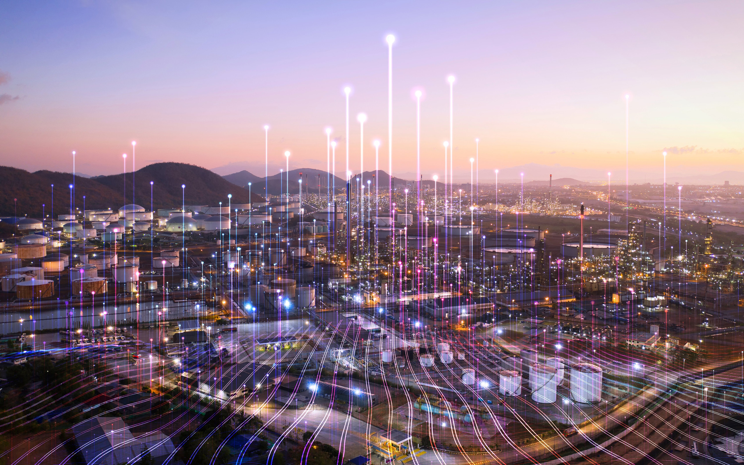 <p>Das Bild zeigt eine weite Aussicht auf eine industrielle Landschaft, wahrscheinlich eine Raffinerie oder ein Produktionsgelände, das am Abend oder in der Dämmerung aufgenommen wurde. Die Szene ist von vielen großen Tanks und Fabrikationsanlagen geprägt, die auf dem Gelände verteilt sind. Über der Landschaft sind zahlreiche vertikale Lichtstrahlen und bunte Linien dargestellt, die eine digitale oder vernetzte Umgebung symbolisieren. Diese visuellen Elemente könnten die Datenübertragung oder Vernetzung durch moderne Technologien darstellen, was auf die digitale Transformation und Smart Manufacturing in der Industrie hinweist. Der Himmel zeigt einen sanften Farbverlauf, der auf den Abendhimmel hinweist.<p>