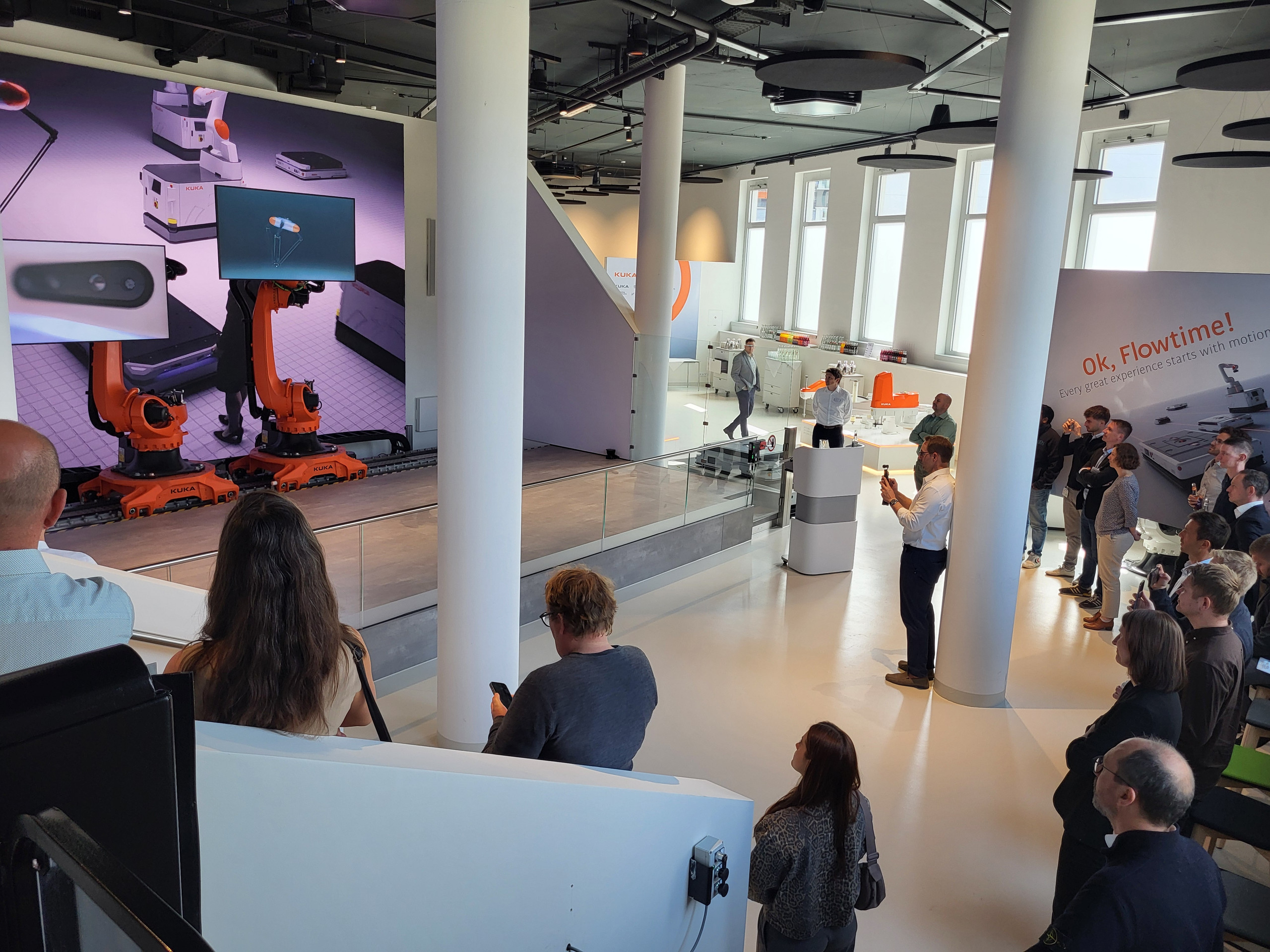<p>Das Bild zeigt: Blick in ein helles Präsentationsforum mit weißen Säulen: Publikum auf Tribüne und im Foyer verfolgt eine Präsentation, große LED‑Wand zeigt KUKA‑Roboter; rechts KUKA‑Branding und Ausstellungsfläche.<p>