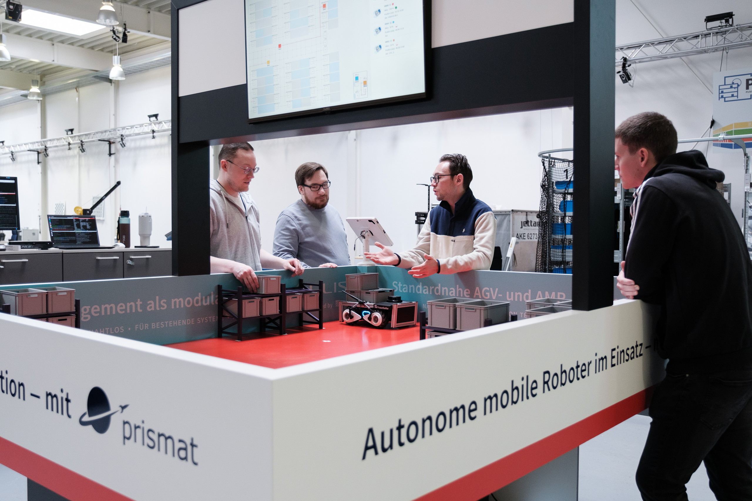 <p>Vier Personen stehen an einem Demonstrationstisch auf einer Messe; ein kleiner autonomer mobiler Roboter bewegt graue Behälter auf rotem Untergrund, darüber hängt ein großer Monitor, an der Booth-Wand steht „Autonome mobile Roboter im Einsatz.<p>